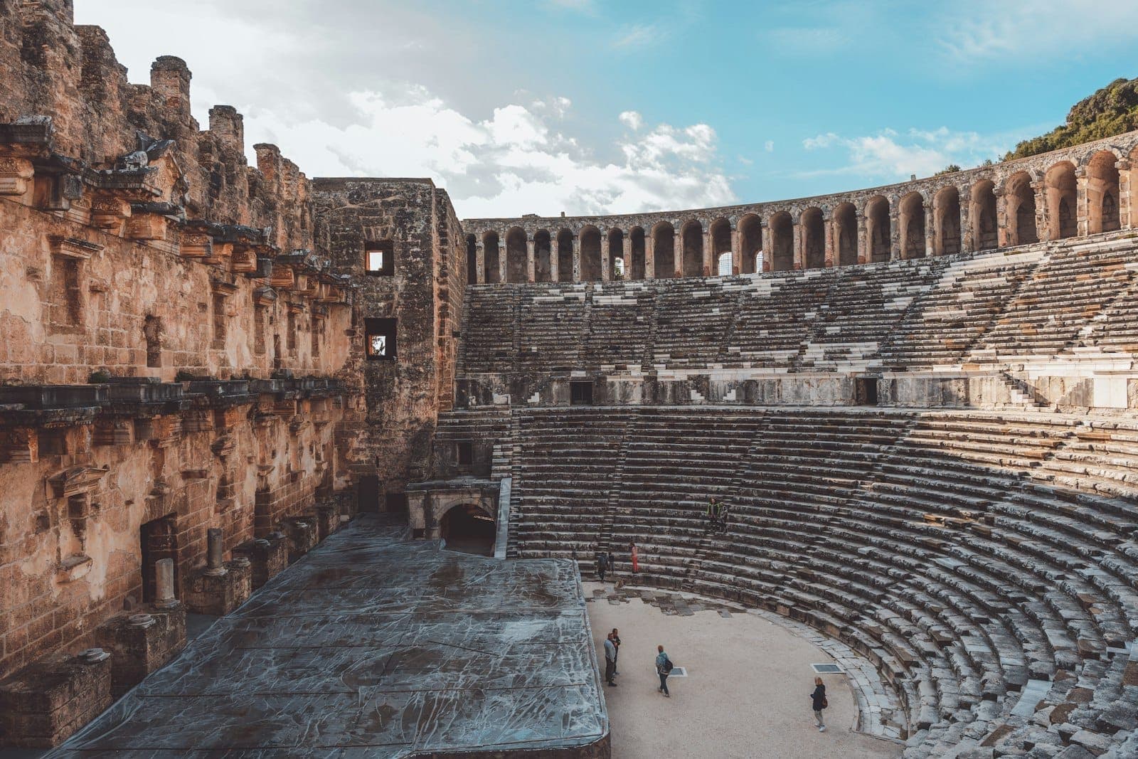 Wide view of the ancient Aspendos Roman theater in Antalya with its stone seating and stage under natural daylight, a few people walking inside.