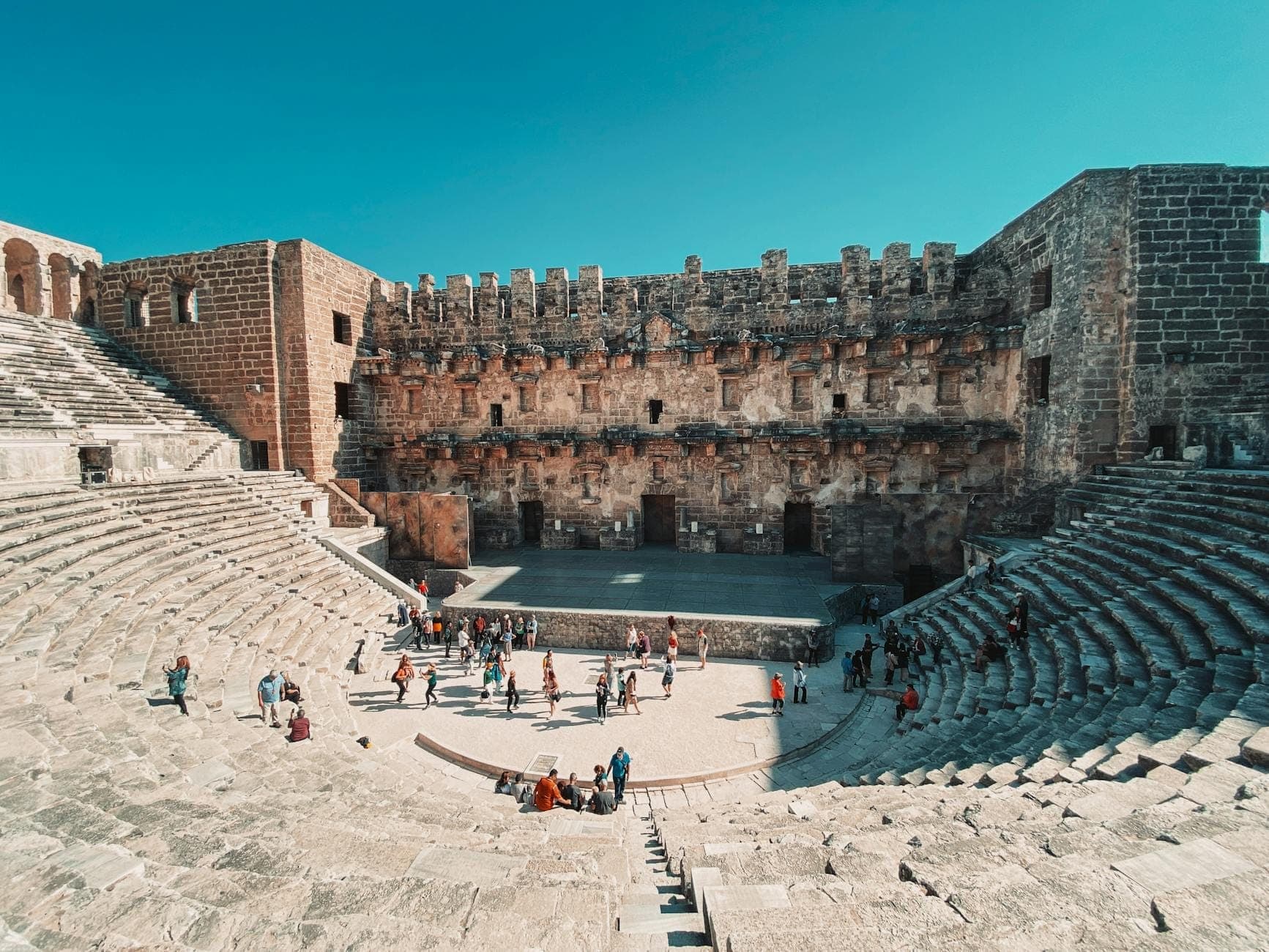 Weitwinkelaufnahme des römischen Theaters von Aspendos unter strahlend blauem Himmel – Besucher erkunden die antiken Steintreppen und die Bühne, ein idealer Ort für Festivals.