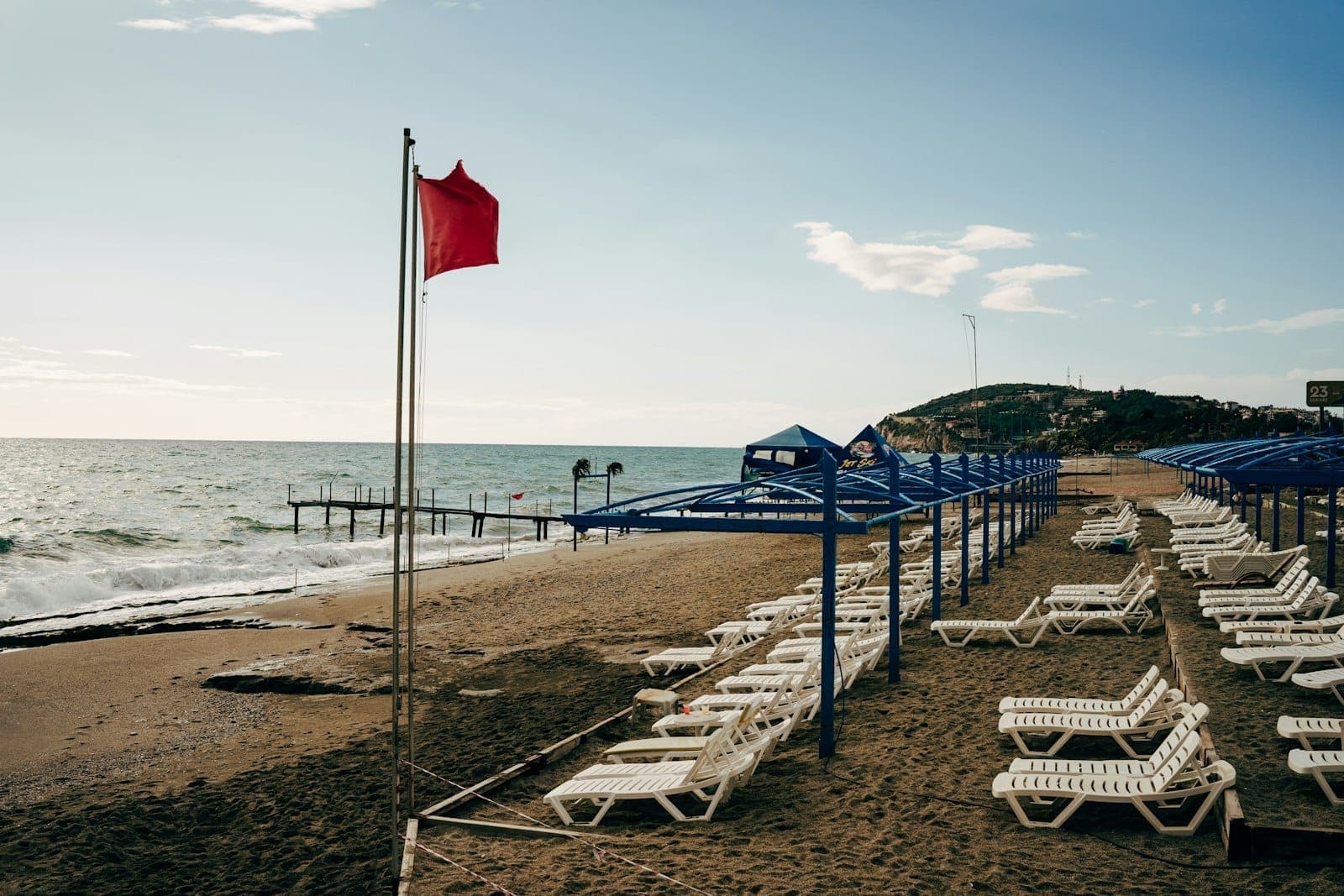 Reihen leerer Sonnenliegen und blauer Sonnenschirme an einem langen Sandstrand mit sanften Wellen und einer roten Flagge, östlich von Antalya.