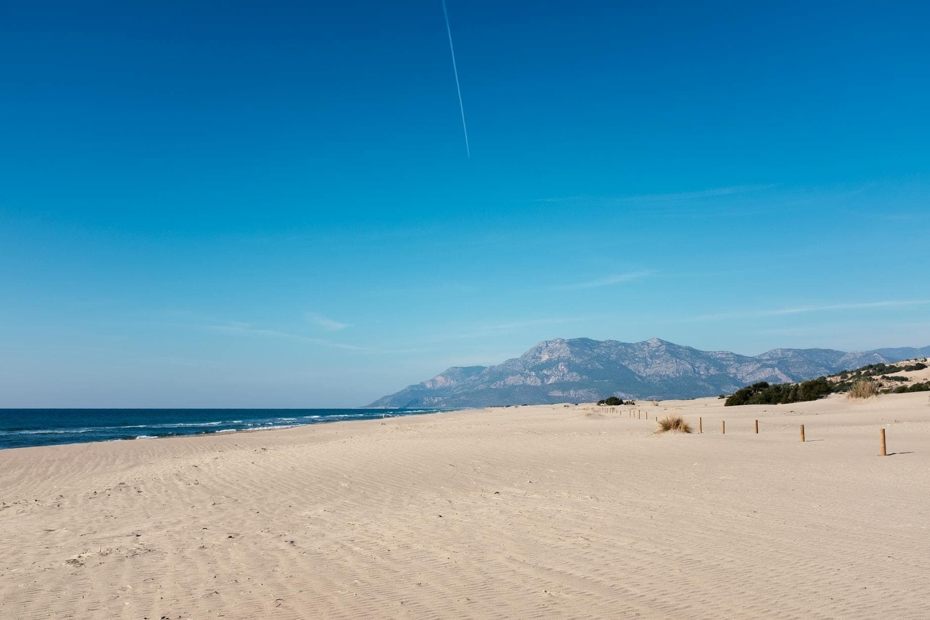 Breiter Sandstrand mit fernen Bergen, blauem Himmel und sanften Wellen; natürliche, offene und einladende Landschaft.