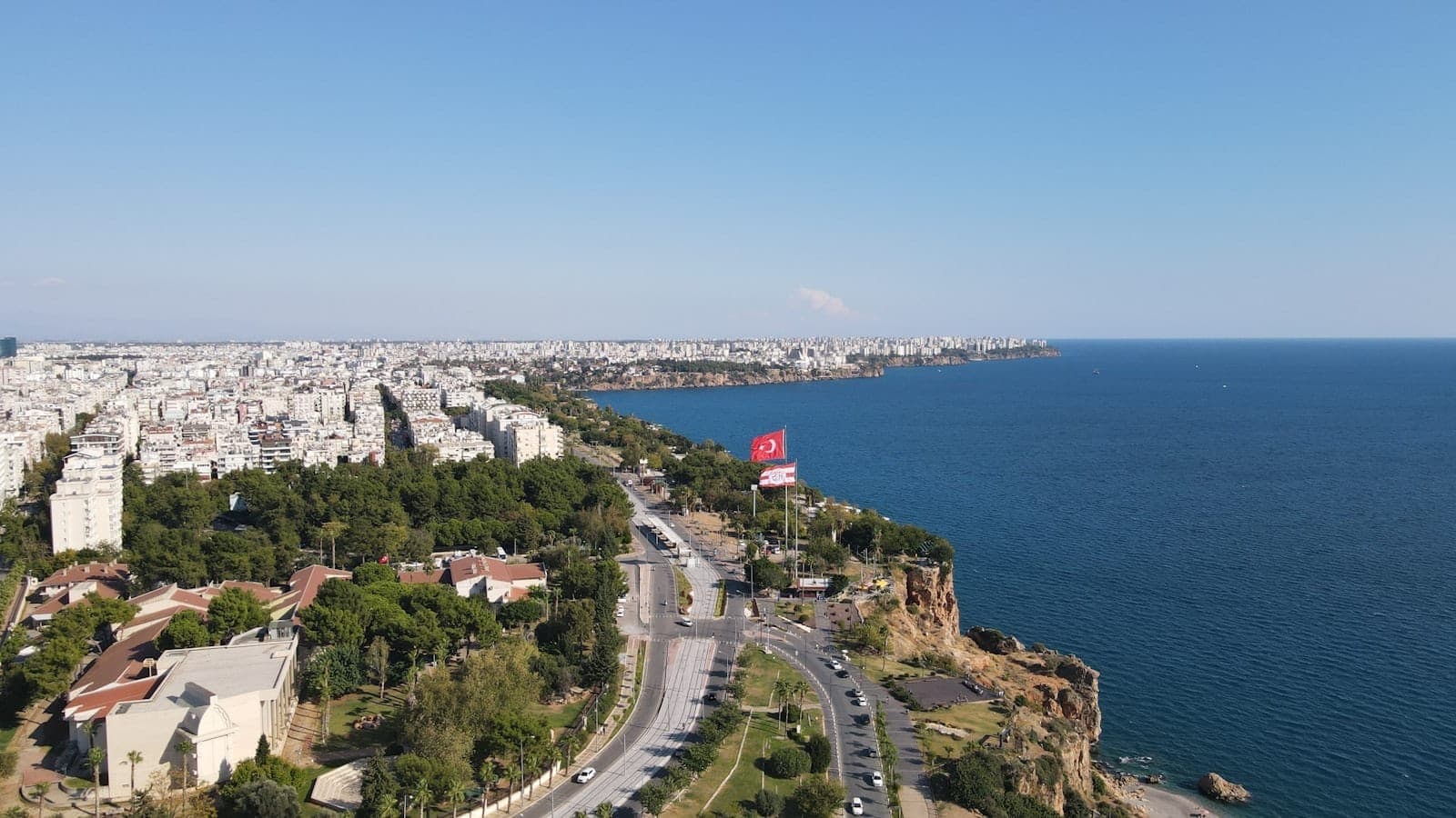 Luftaufnahme von Antalya mit Küstenlinie, Stadtsilhouette, breiten Straßen, Grünflächen und einer großen türkischen Flagge unter strahlend blauem Himmel.