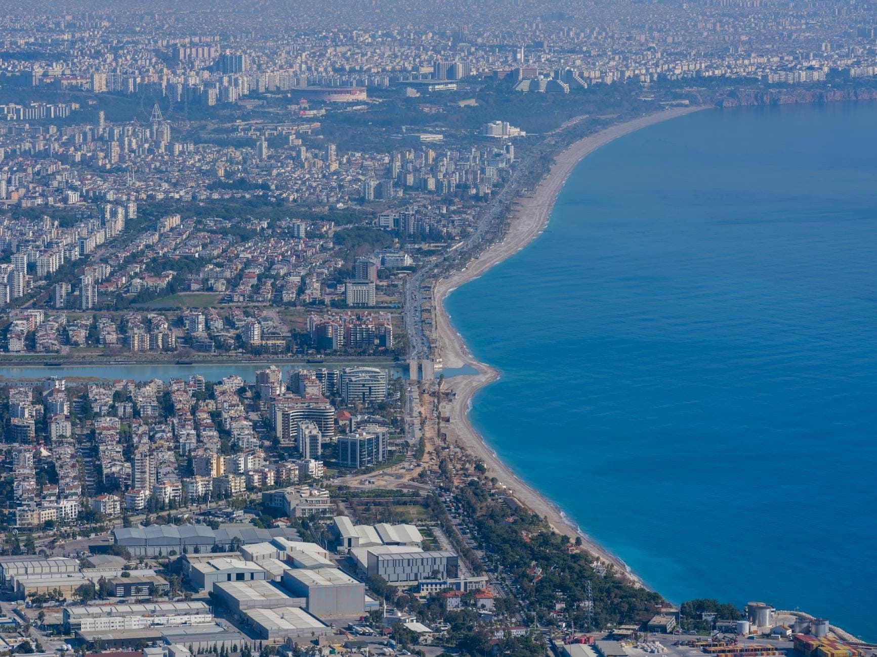 Luftaufnahme von Antalya mit der weitläufigen Stadt, der Küstenlinie und dem Bogen des Konyaaltı-Strandes, der auf das türkisfarbene Mittelmeer trifft.