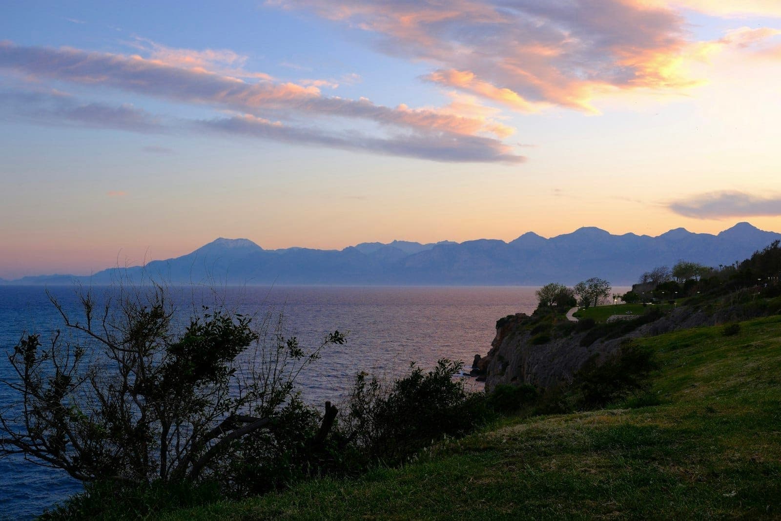 Klippenpark in Antalya bei Sonnenuntergang mit Meerblick, Baumsilhouetten und fernen Bergen unter einem farbenfrohen Himmel.