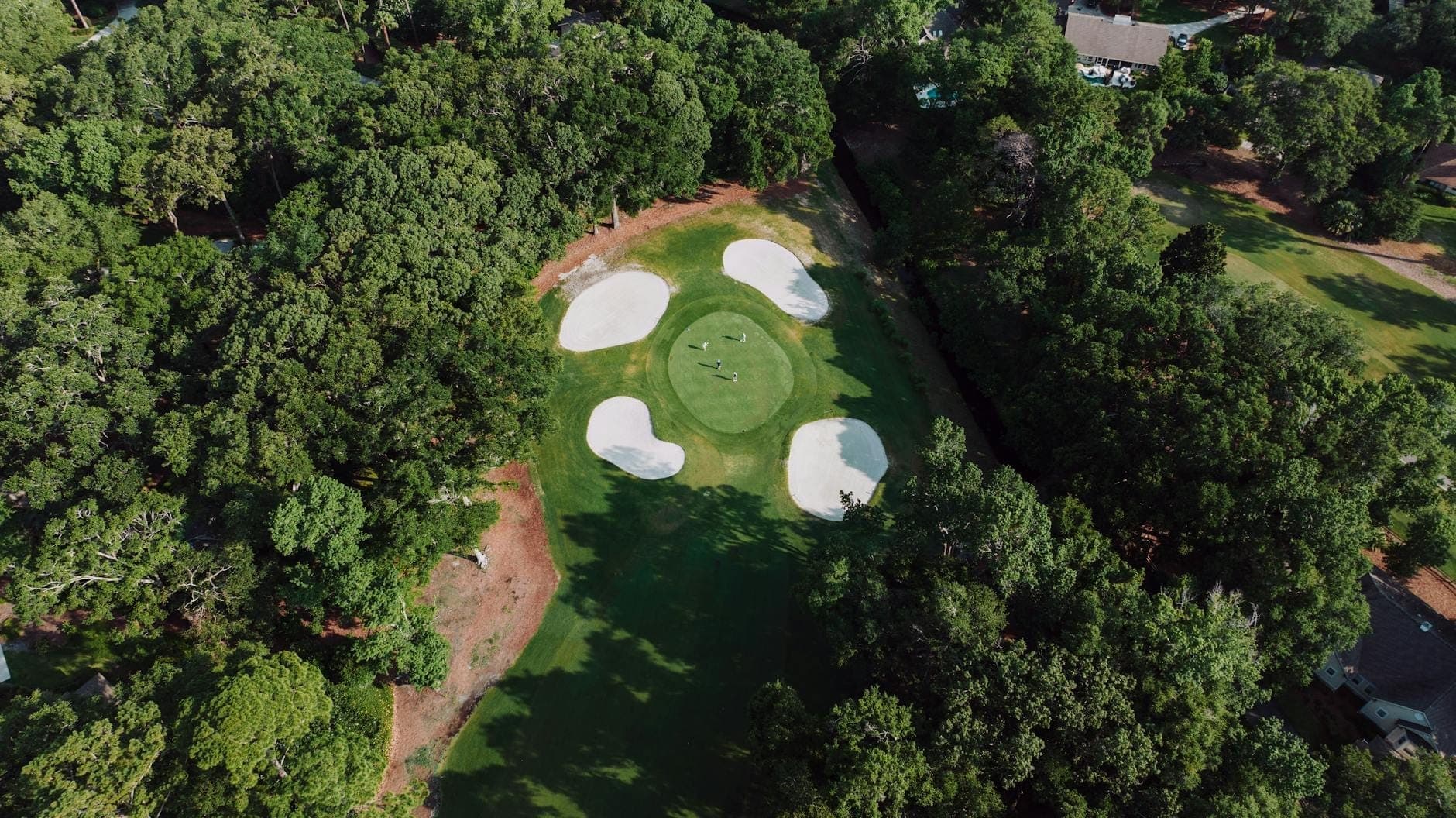 Luftaufnahme eines grünen Golfplatzes, umgeben von Bäumen mit sichtbaren Sandbunkern und gepflegten Fairways.
