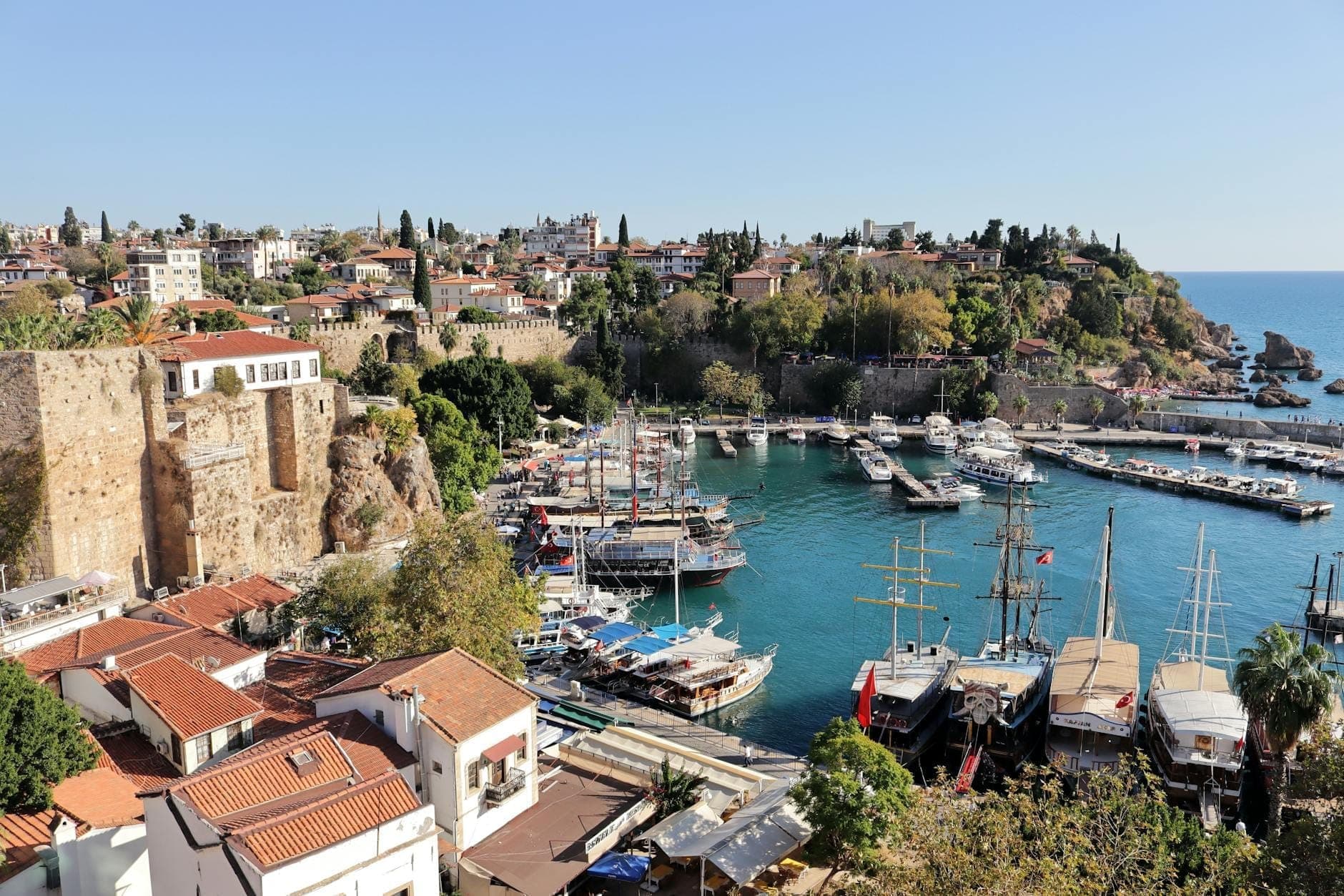 Le vieux port d'Antalya par une belle journée ensoleillée, bateaux en bois traditionnels sur une eau turquoise, remparts historiques en pierre et maisons aux toits rouges sous un ciel d'été dégagé.