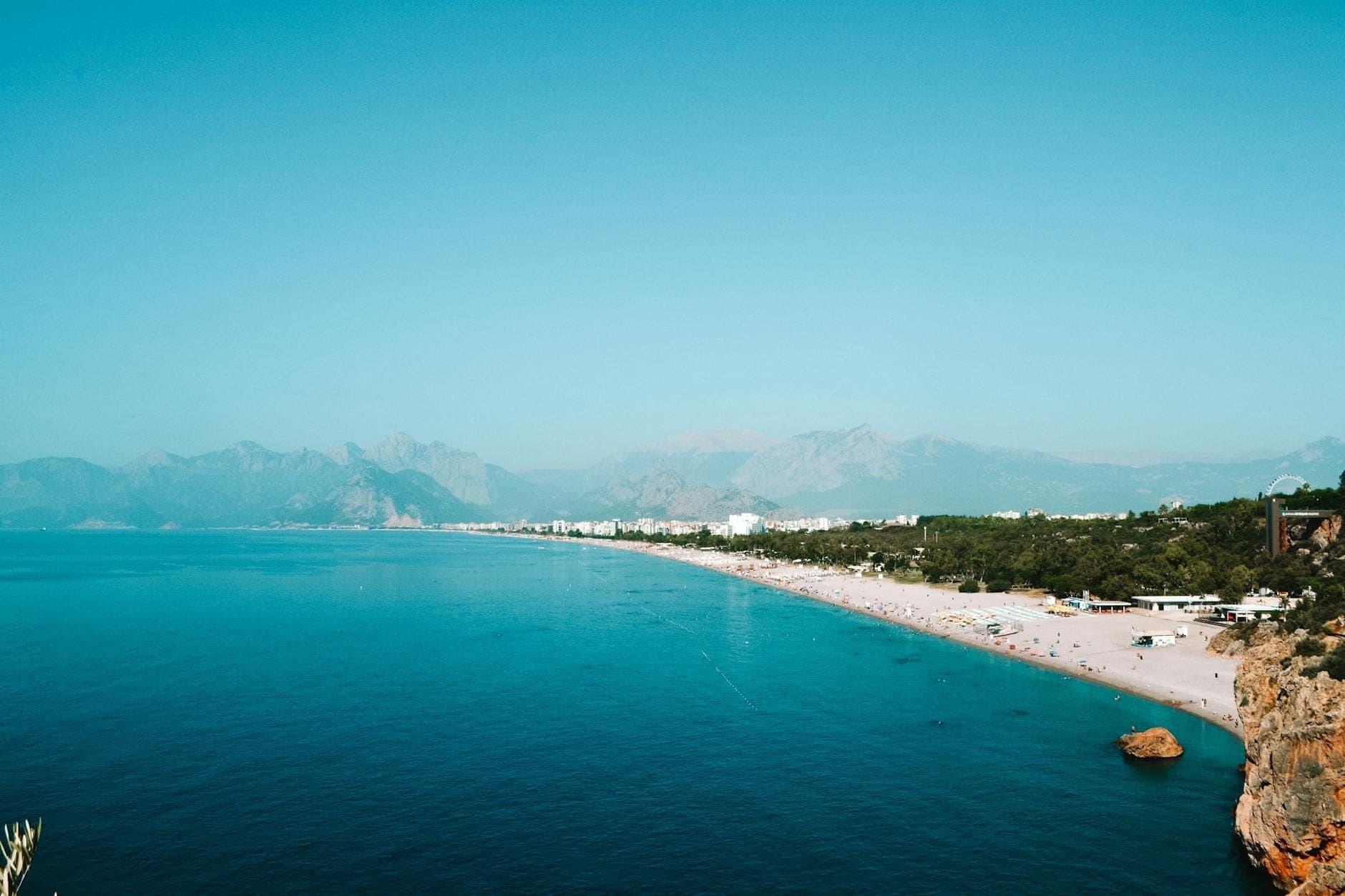 Vue panoramique de la plage de Konyaaltı à Antalya avec une mer turquoise, un rivage sablonneux, des montagnes au loin et des baigneurs épars, sous un ciel bleu dégagé.