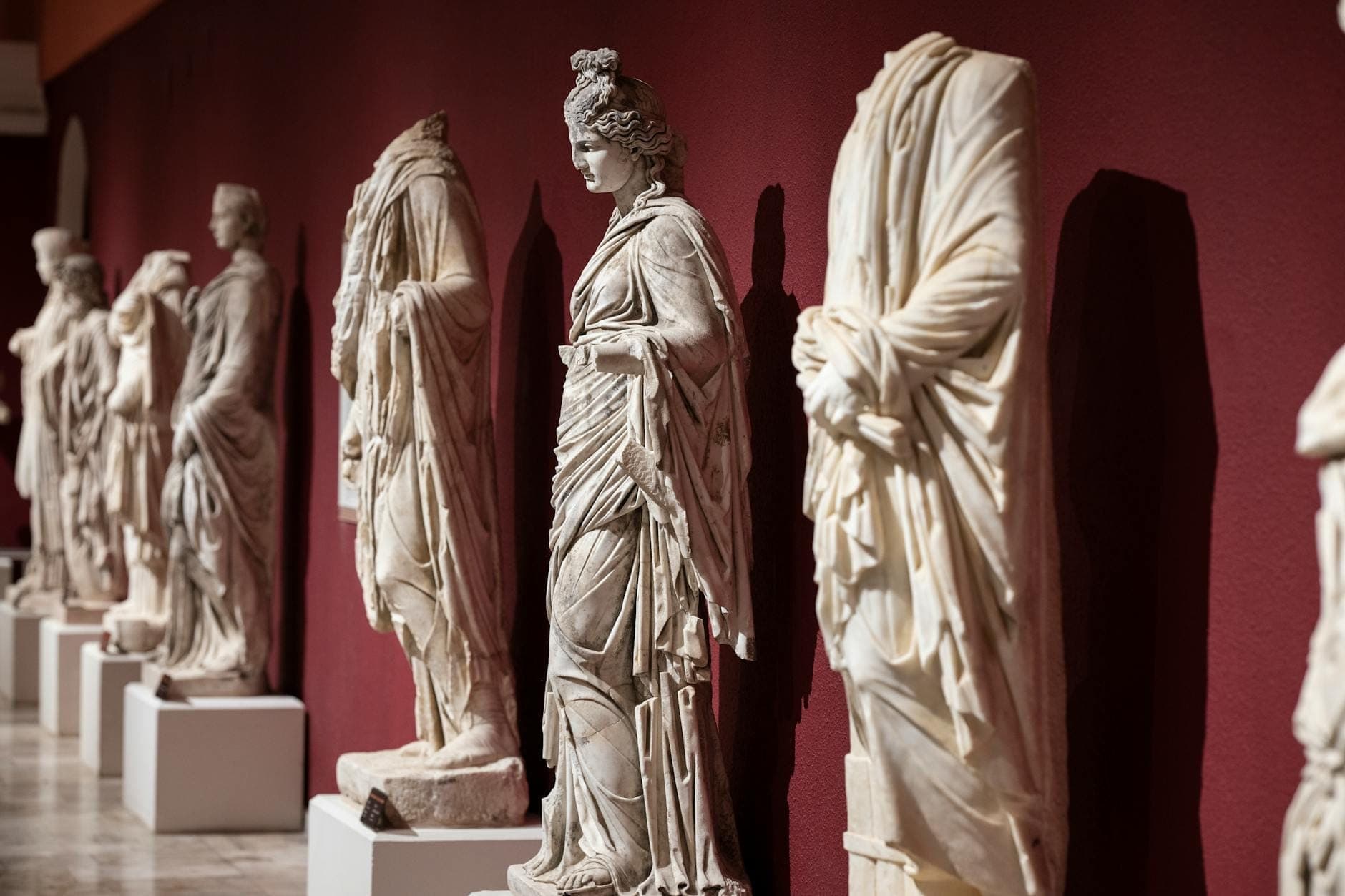 Eine Reihe antiker Marmorstatuen aus der Römerzeit, ausgestellt in einem Innenraum vor einer roten Wand im Archäologischen Museum Antalya.