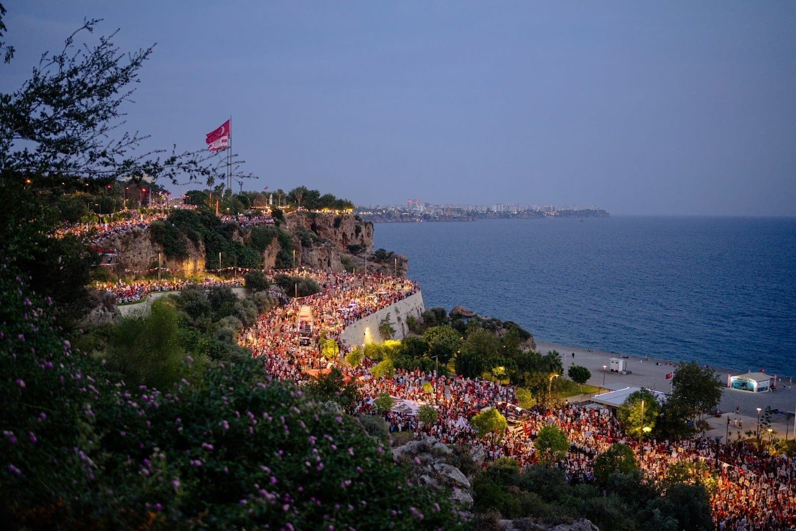 Des foules rassemblées le long des spectaculaires falaises côtières d'Antalya au crépuscule, avec des lumières vives et une atmosphère estivale festive au bord de l'eau.