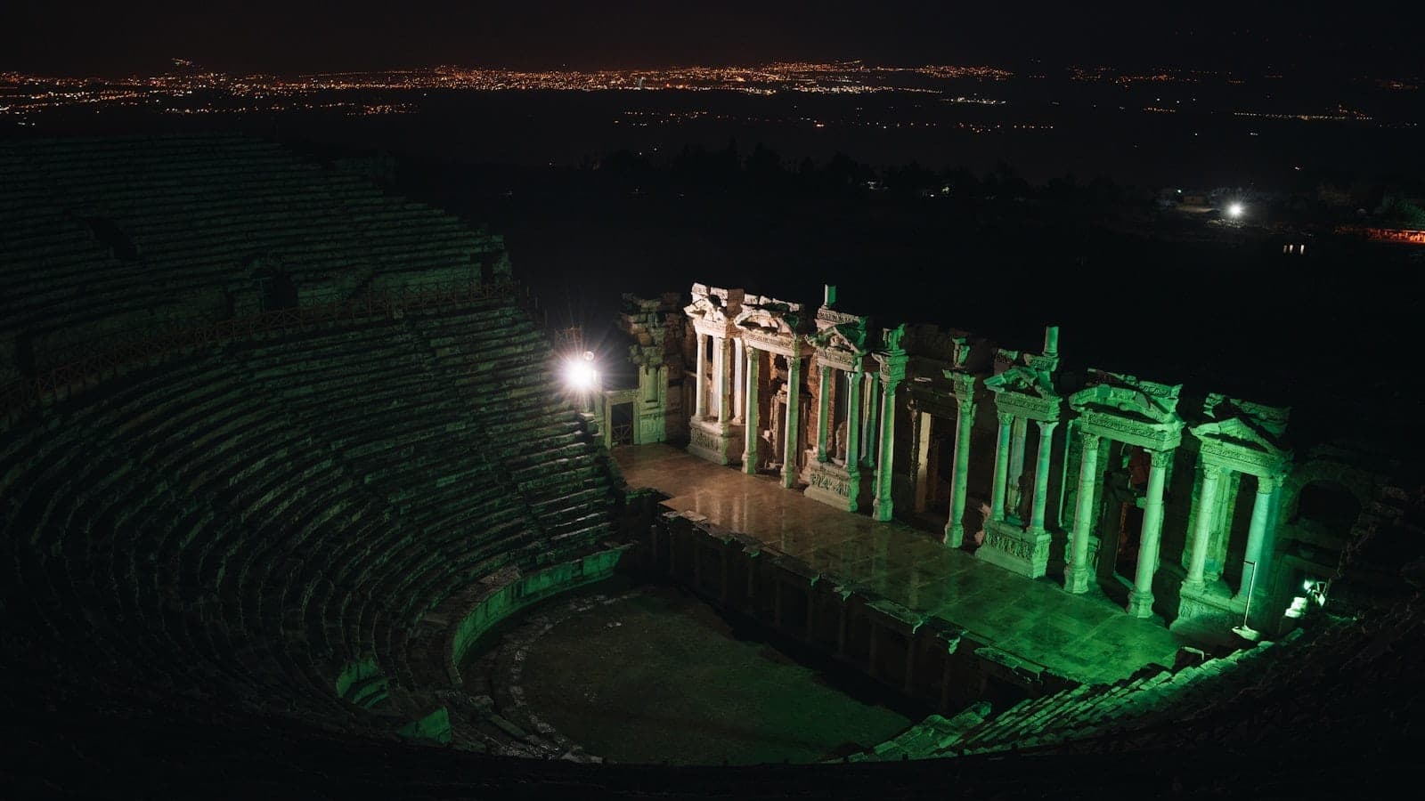 Vue nocturne d'un grand amphithéâtre romain vivement illuminé, avec les lumières de la ville en arrière-plan, évoquant un événement culturel en soirée.