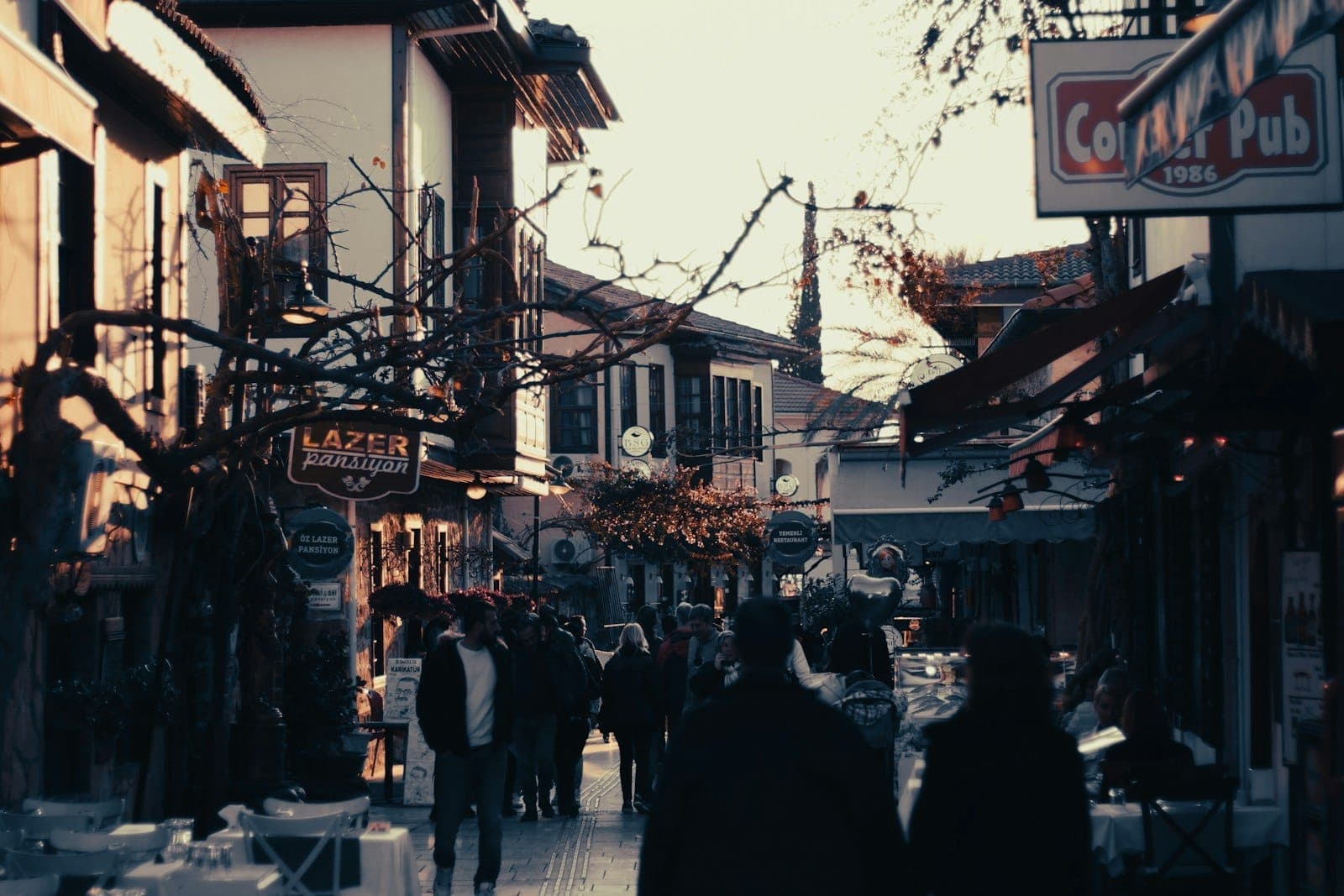 Rue de la vieille ville d'Antalya remplie de personnes, bars, cafés et enseignes dans la lumière du début de soirée.