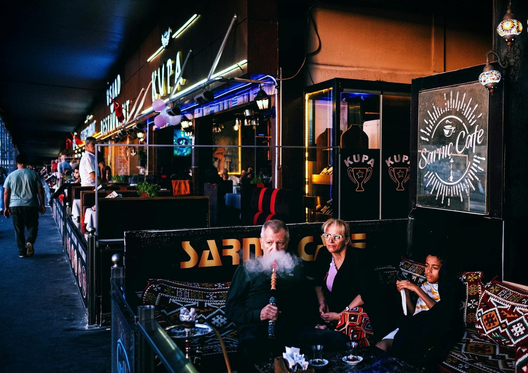 Des personnes savourant le shisha dans un bar en plein air à l'atmosphère unique avec des néons et des tapis turcs, capturant la vie nocturne authentique à Antalya.