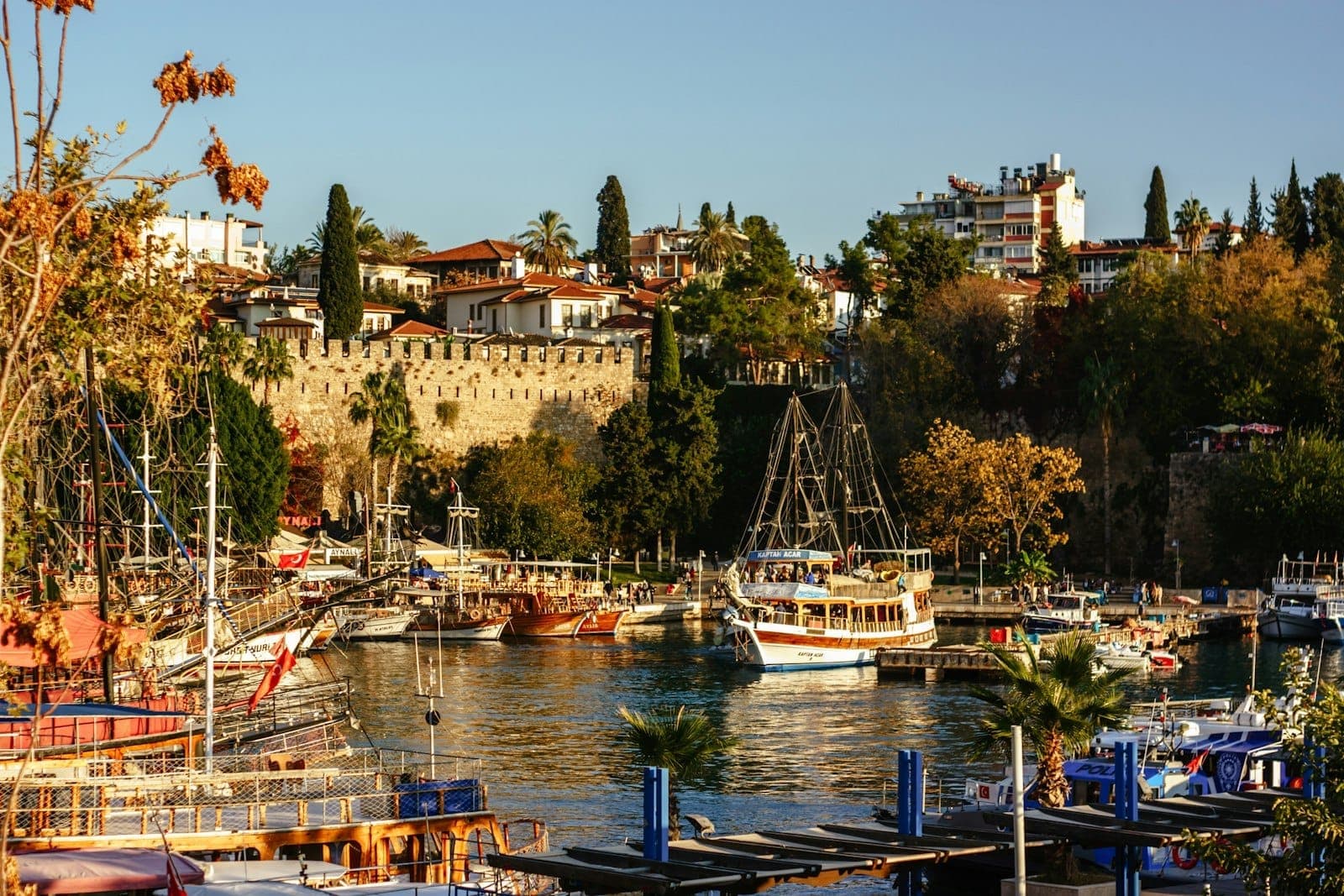Bunte Boote im historischen Hafen von Antalya, im Hintergrund alte Stadtmauern, üppige Bäume und Wohngebäude an einem sonnigen Tag.