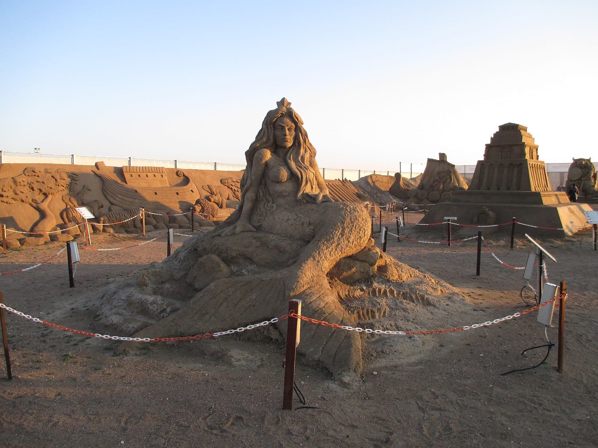 Große Sandskulpturen, darunter eine Meerjungfrau und antike Bauwerke, ausgestellt im Sandland Antalya unter einem klaren Abendhimmel.