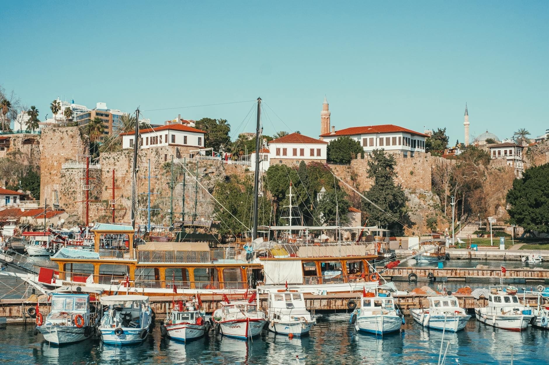 Vue du port d'Antalya avec des bateaux amarrés devant la vieille ville et des bâtiments historiques en pierre sous un ciel bleu dégagé.