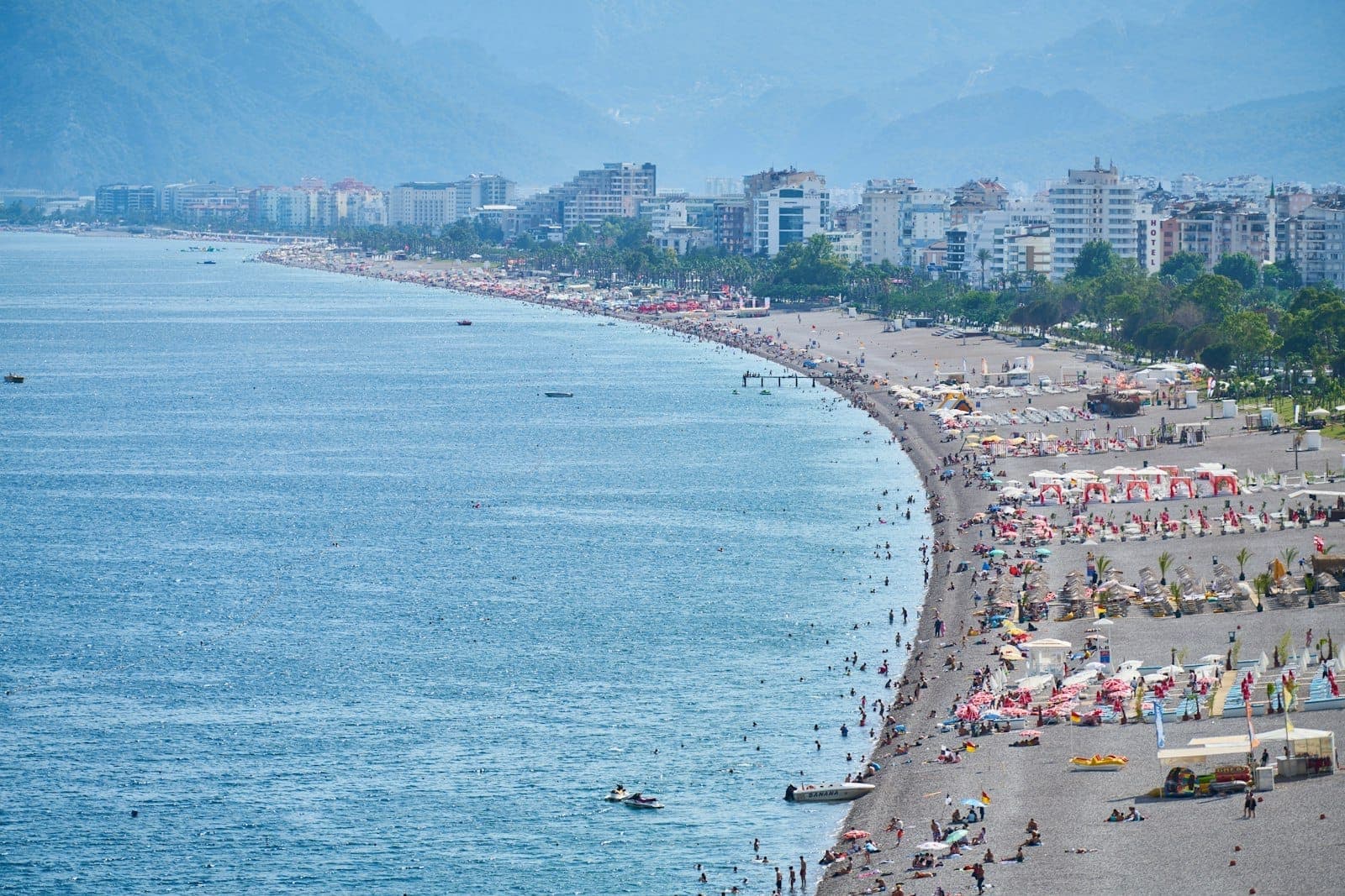 Vue large de la plage urbaine d'Antalya bondée de personnes, de parasols et de gratte-ciels en pleine saison estivale.