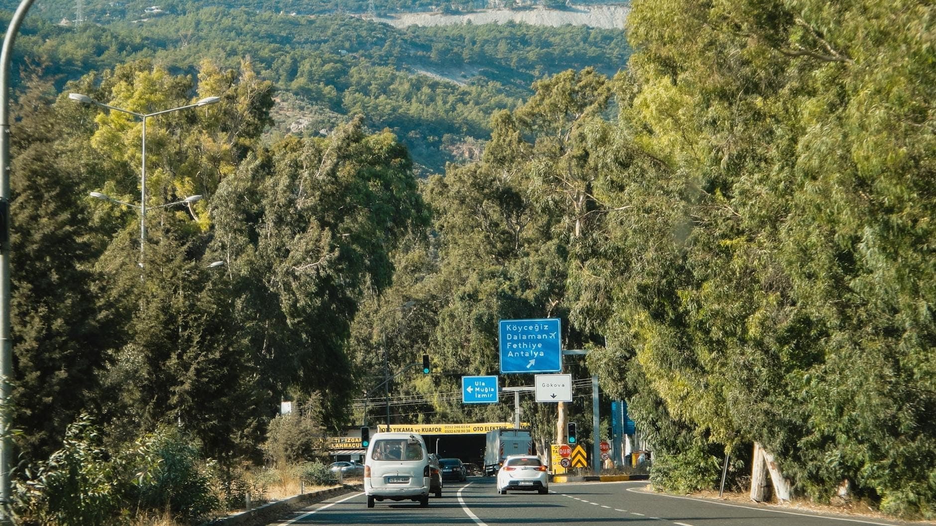 View of cars driving on a tree-lined highway near Antalya, with road signs and mountains in the background.