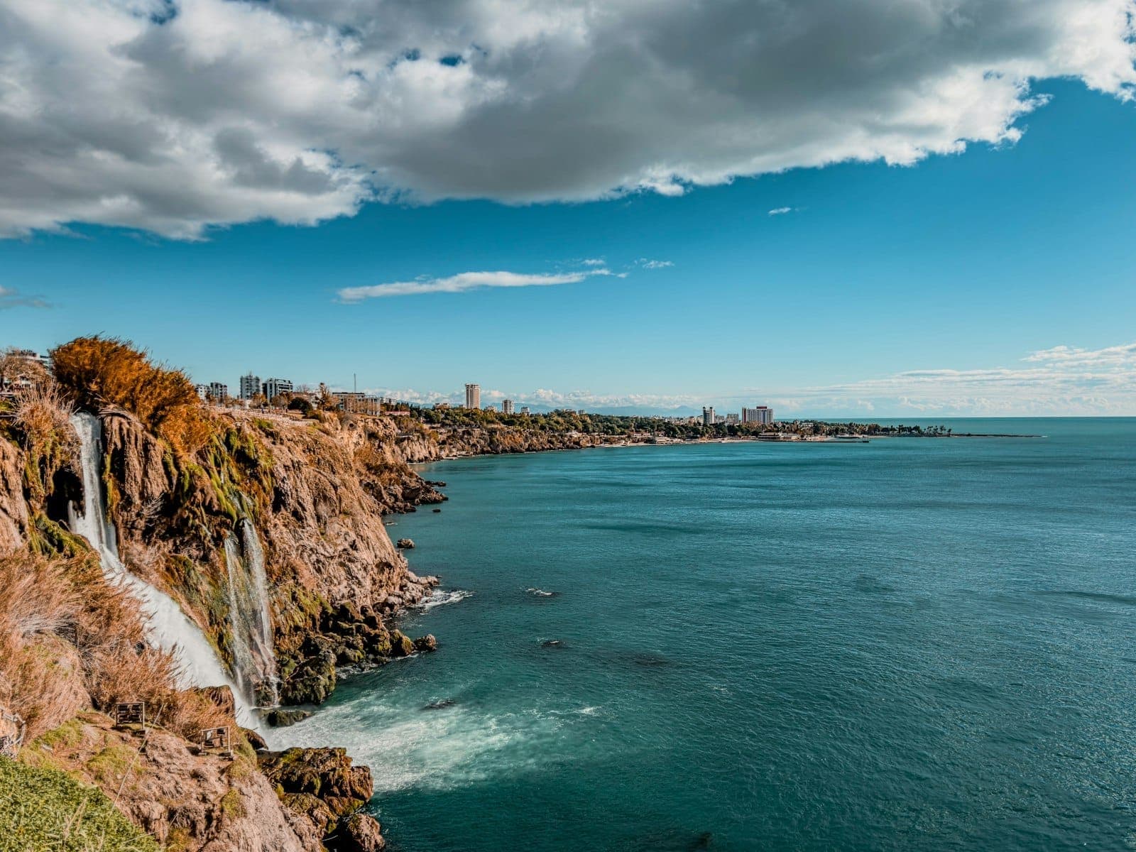 Weitwinkelansicht der unteren Düden-Wasserfälle bei Antalya, die ins tiefblaue Mittelmeer stürzen – mit Stadtsilhouette und dramatischen Wolken im Hintergrund.