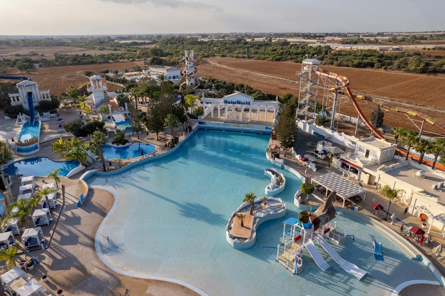 Vue aérienne d'un grand parc aquatique avec piscines, toboggans et aires de jeux, entouré de chaises longues et de palmiers, sous un ciel partiellement nuageux.