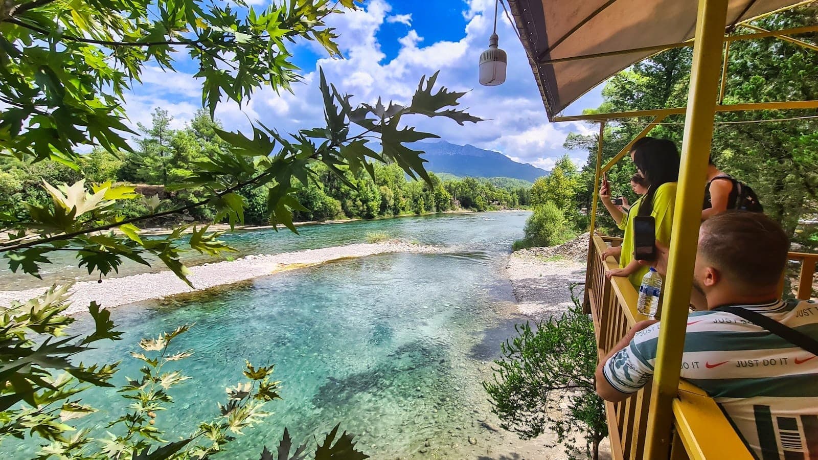 Kinder und Erwachsene genießen eine malerische Flussterrasse mit kristallklarem Wasser, grünen Bäumen und fernen Bergen unter strahlendem Himmel bei Antalya.