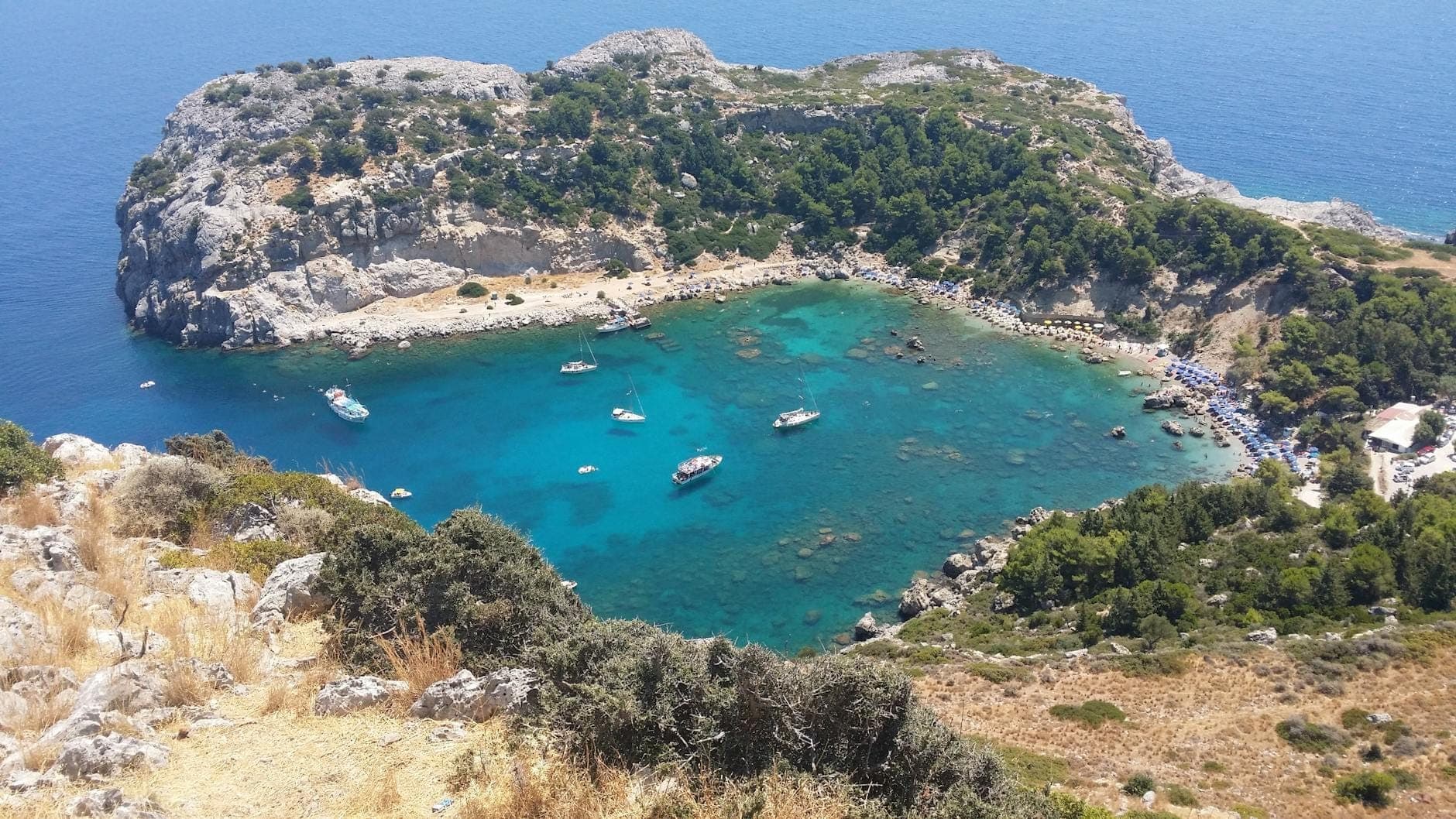 Vue aérienne de la baie d'Anthony Quinn avec son eau turquoise, des bateaux ancrés, des falaises rocheuses et une végétation verdoyante longeant le rivage par une journée ensoleillée.