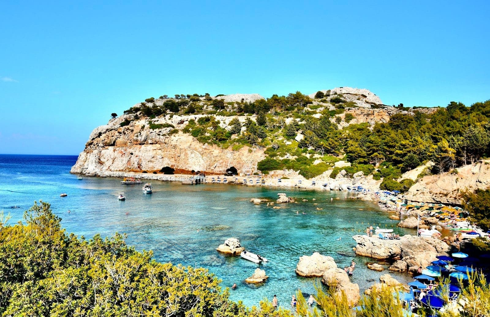 La baie Anthony Quinn à Rhodes avec une eau turquoise cristalline, des falaises rocheuses, des pins, des baigneurs et des bateaux amarrés dans la crique abritée.