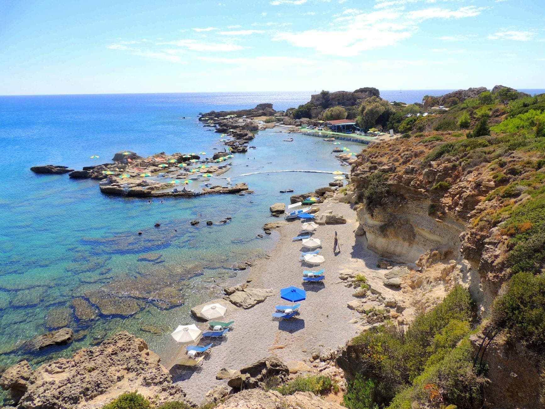 Skalista zatoczka z krystalicznie czystą turkusową wodą, rozstawionymi parasolami, leżakami i otaczającą zielenią w Anthony Quinn Bay na Rodos.