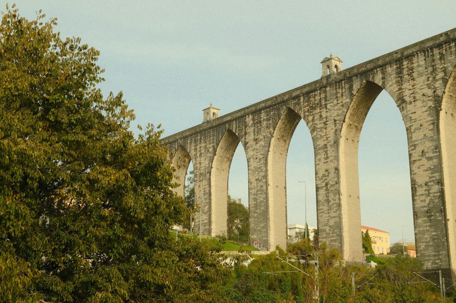 Vista do Aqueduto das Águas Livres em Lisboa, com os altos arcos de pedra góticos acima das copas das árvores e o céu azul ao fundo.