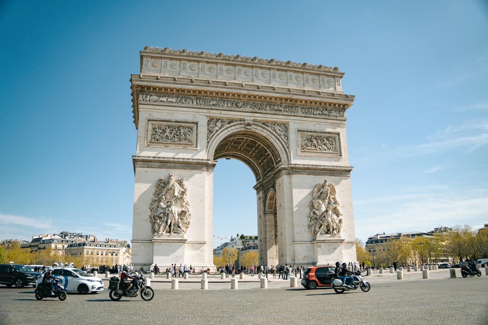 Pemandangan sudut lebar Arc de Triomphe di siang hari, dengan langit biru, orang-orang, dan kendaraan di sekitar monumen. Gedung-gedung dan pepohonan khas Paris terlihat di latar belakang.