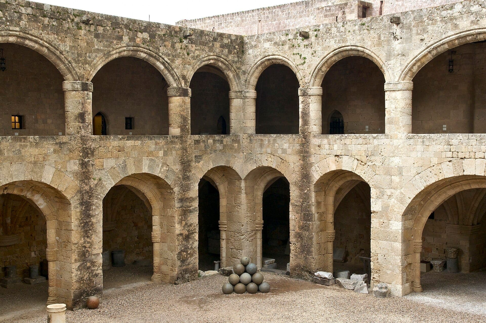 Arches en pierre et cour médiévale du Musée Archéologique de Rhodes, avec des boulets de canon empilés au centre sur un sol en gravier.