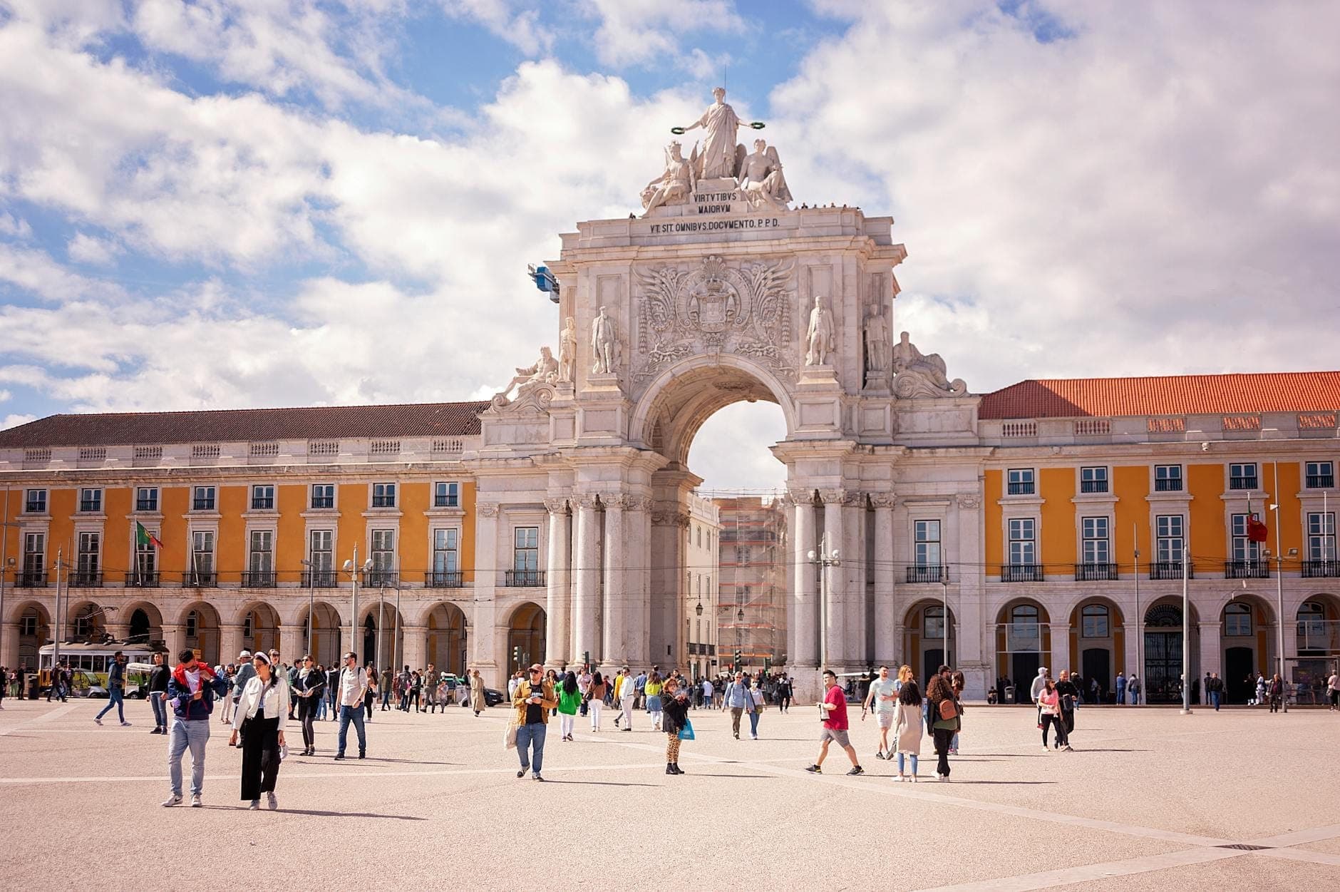 Vista animada e luminosa do Arco da Rua Augusta com multidões de turistas na Praça do Comércio, prédios amarelos e um céu azul com nuvens esparsas.