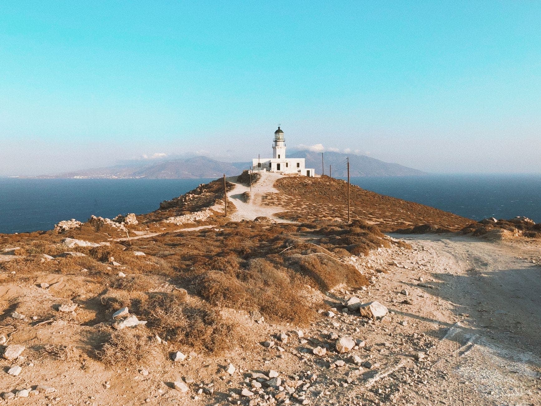 Der Leuchtturm Armenistis auf einem felsigen Hügel mit Blick auf die blaue Ägäis, ein gewundener Weg führt hinauf, im Hintergrund entfernte Berge.