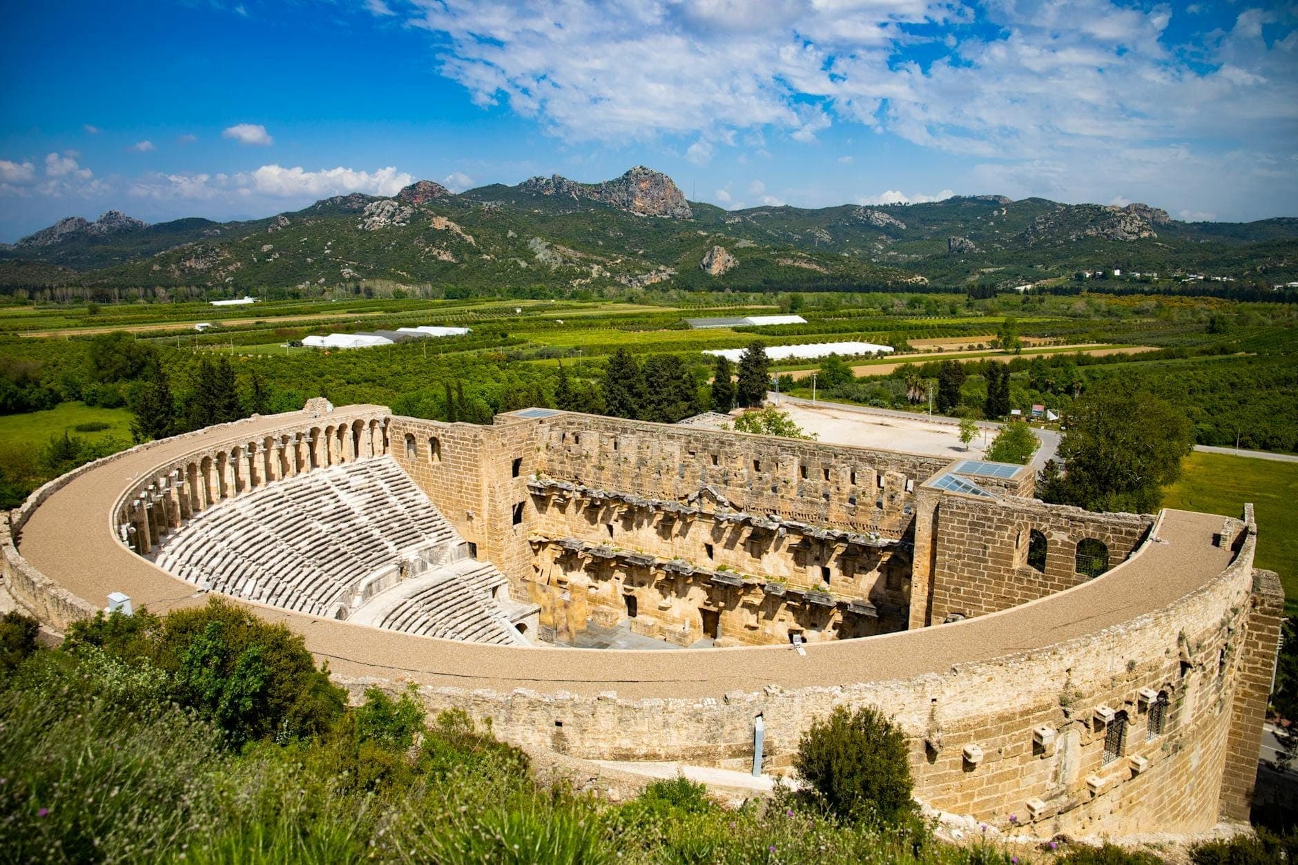 Weitwinkelpanorama des antiken Aspendos Theaters, umgeben von satten grünen Feldern und fernen Bergen unter einem strahlend blauen Himmel mit vereinzelten Wolken.