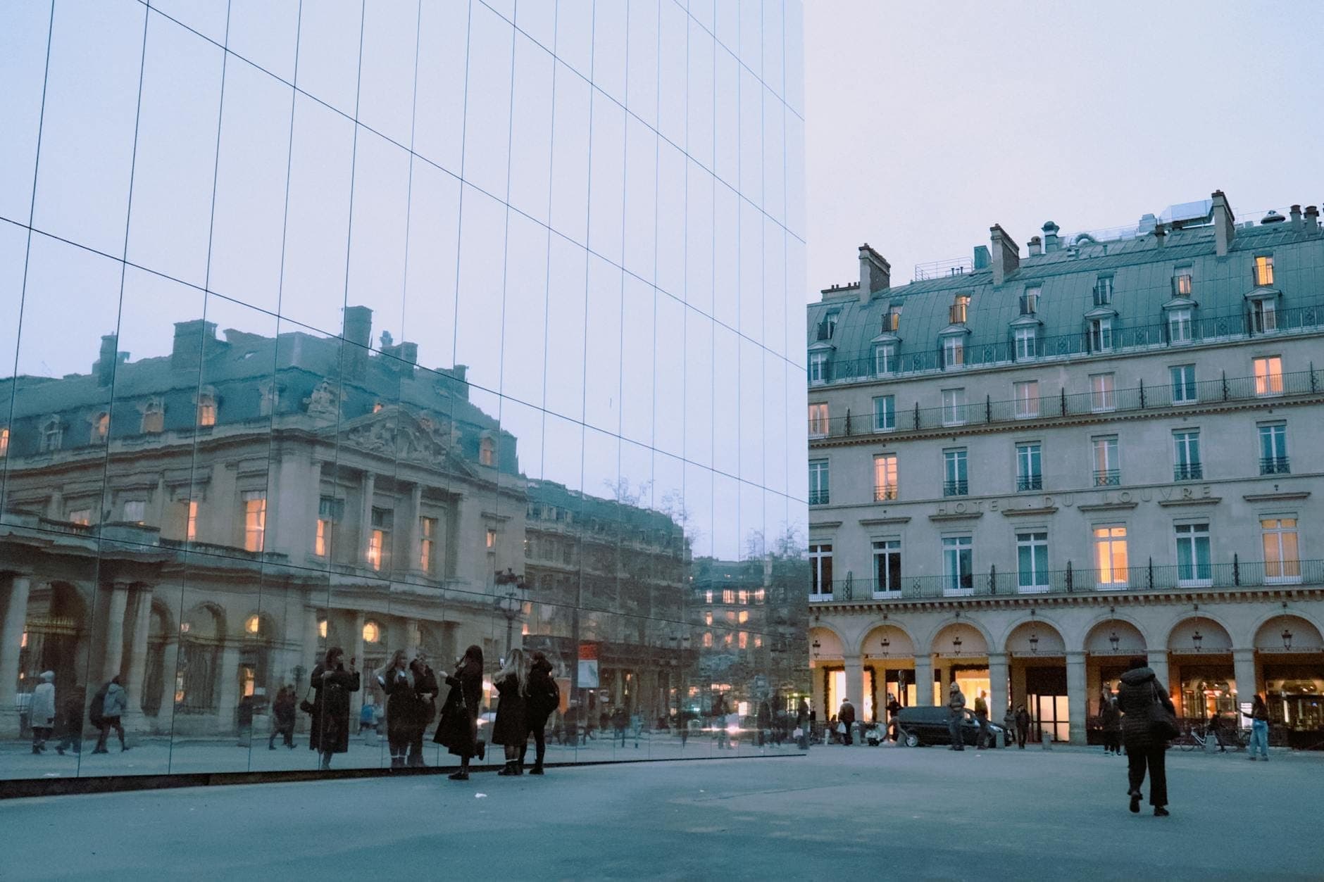 Modern glass building reflecting historic Parisian architecture with people walking in the foreground during dusk.