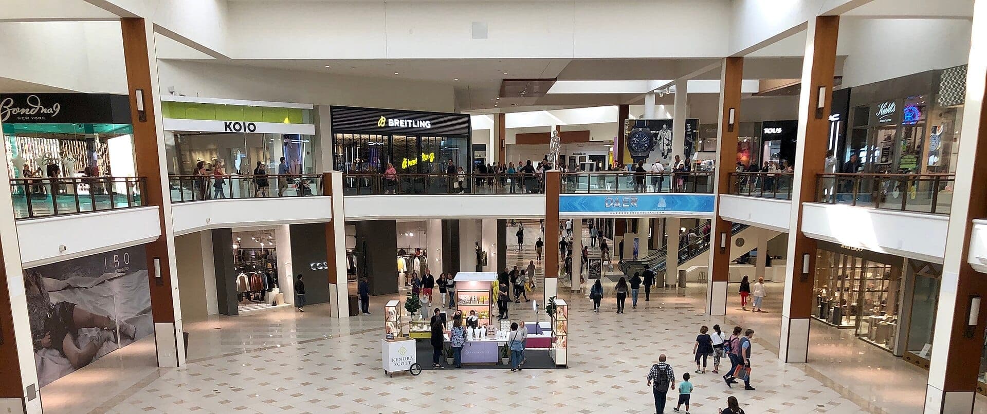 Vue intérieure spacieuse de l'Aventura Mall avec plusieurs boutiques de luxe, des clients se promenant et une atmosphère moderne et lumineuse avec un éclairage naturel.