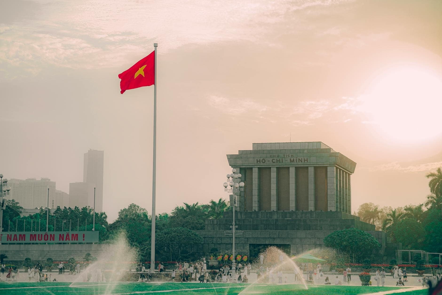 Plac Ba Dinh o zachodzie słońca z wysoko powiewającą wietnamską flagą, fontannami na pierwszym planie i wyraźnie widocznym Mauzoleum Ho Chi Minha.