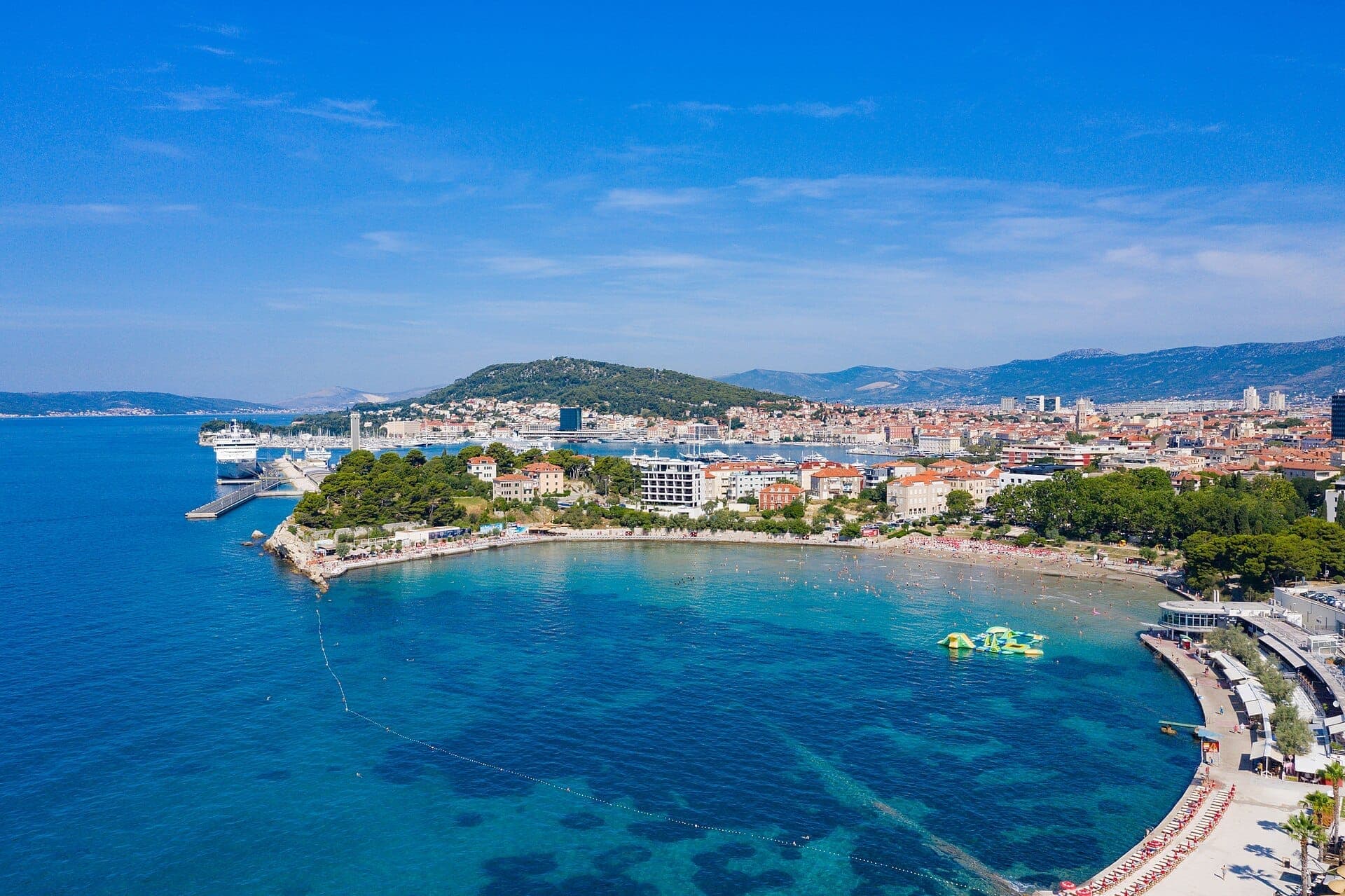 Vue aérienne du rivage en croissant de la plage Bačvice, eau turquoise claire, bâtiments en bord de mer et la vieille ville de Split et ses collines en arrière-plan.