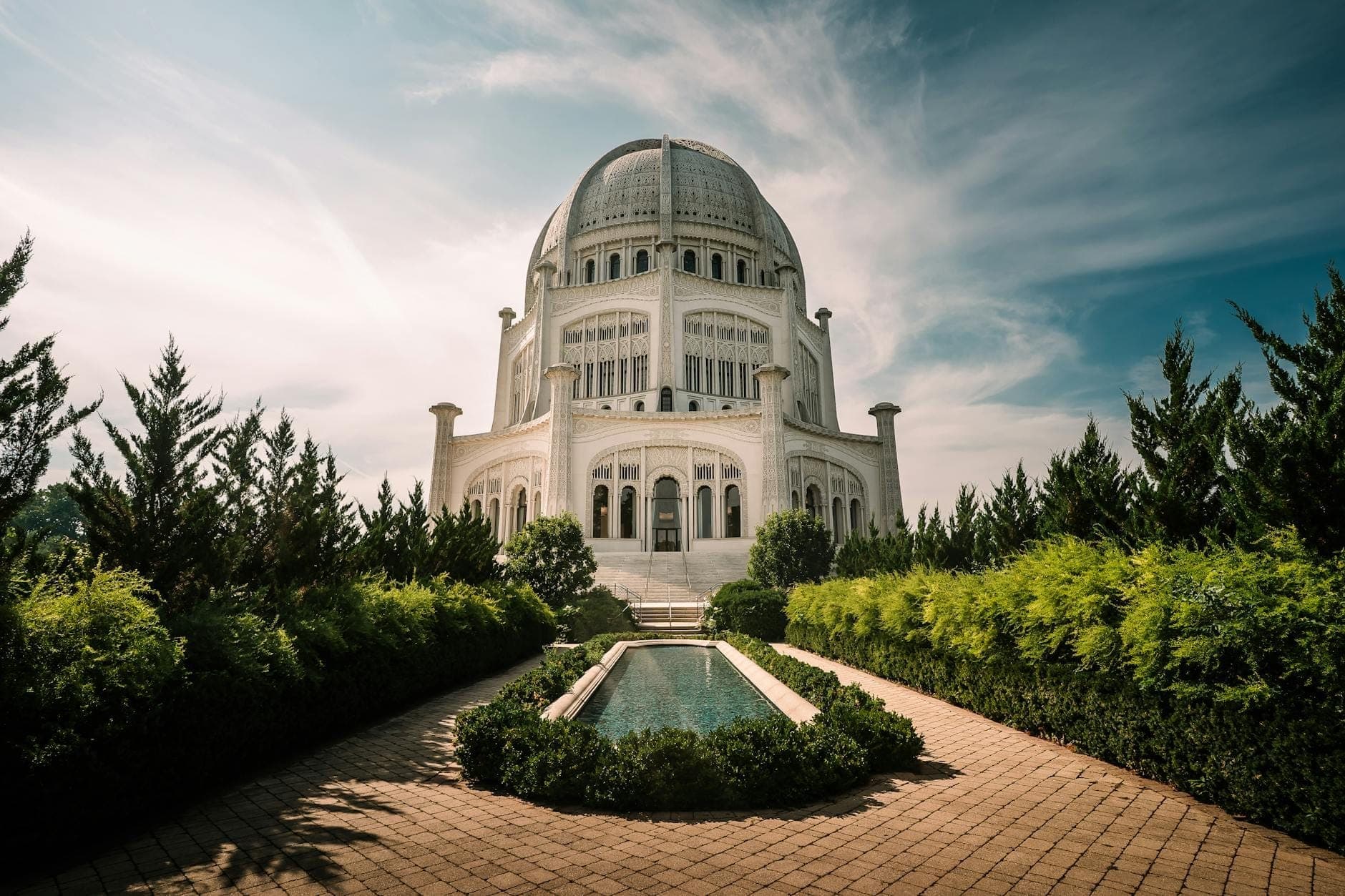 Vista panorámica del Templo Bahá'í en Wilmette, enmarcado por vegetación exuberante y un estanque reflectante, bajo un cielo azul brillante con nubes ligeras.