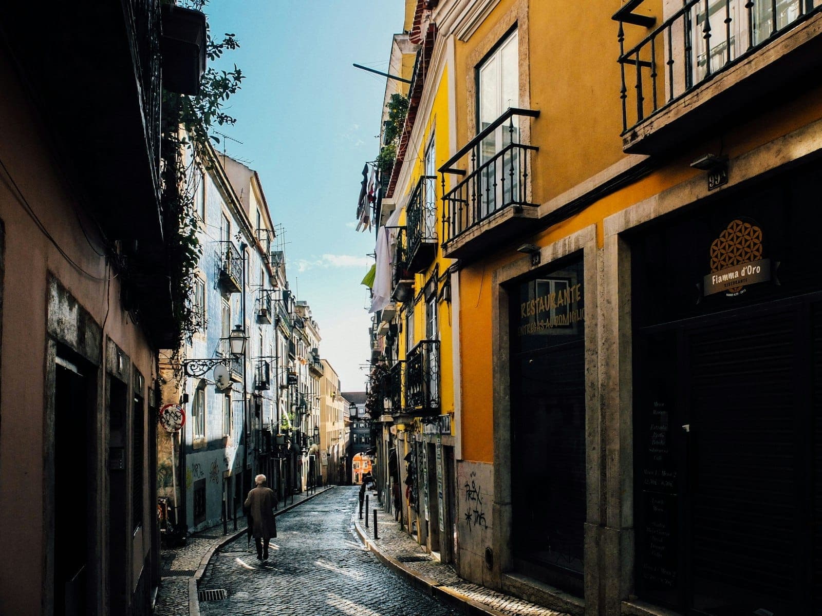 Rua estreita de paralelepípedo no Bairro Alto, ladeada por prédios antigos e coloridos com varandas, com algumas pessoas caminhando à luz do dia.