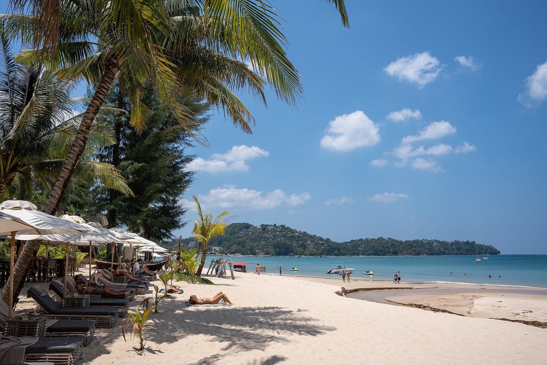 People relax on sun loungers under palm trees at Bang Tao Beach, with soft white sand, clear blue water, and distant green hills.