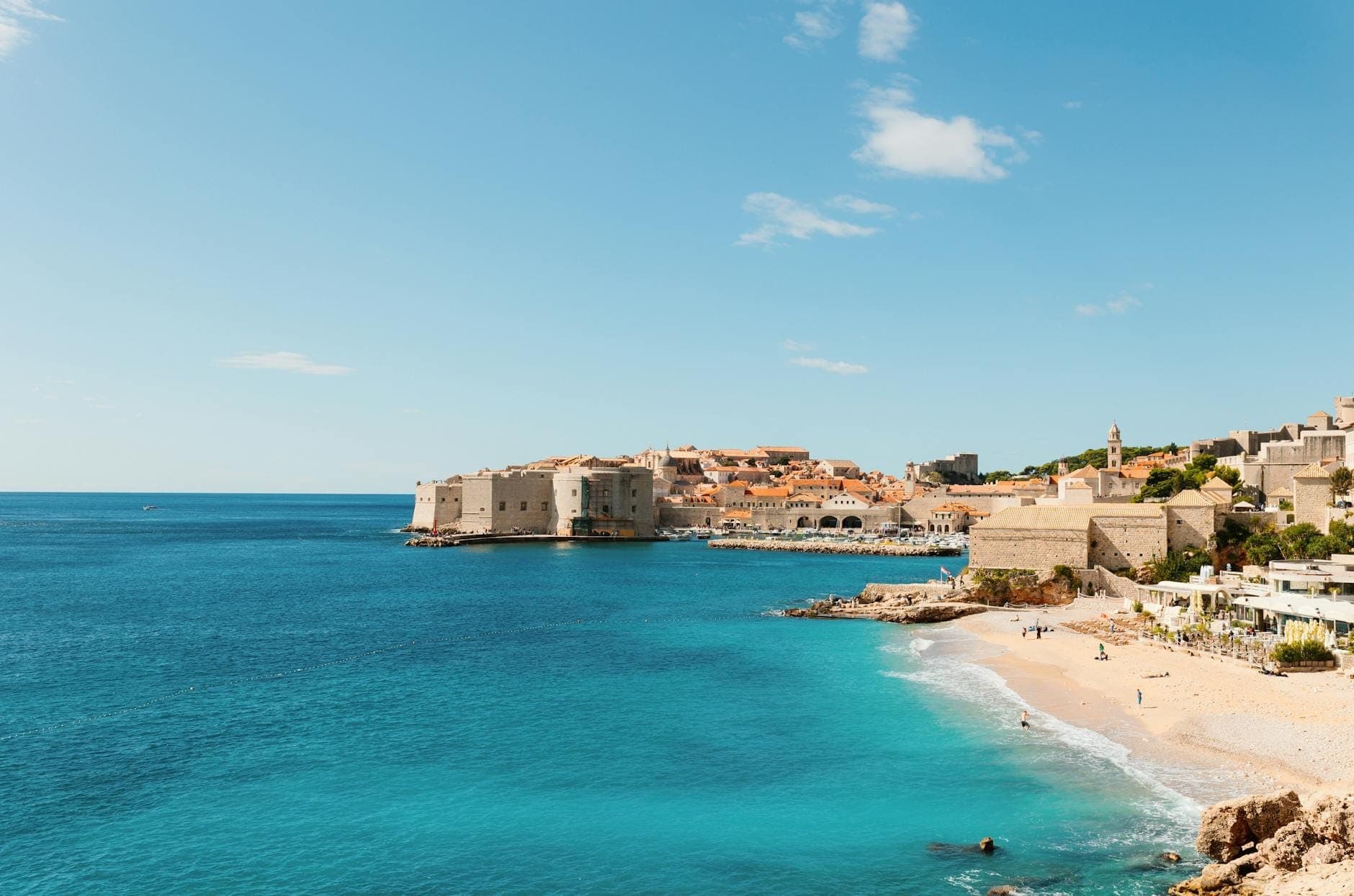 Beeindruckender Blick auf die Banje Beach mit ihrem Kieselstrand, dem klaren blauen Wasser und den mittelalterlichen Mauern der Dubrovniker Altstadt im Hintergrund unter strahlend blauem Himmel.