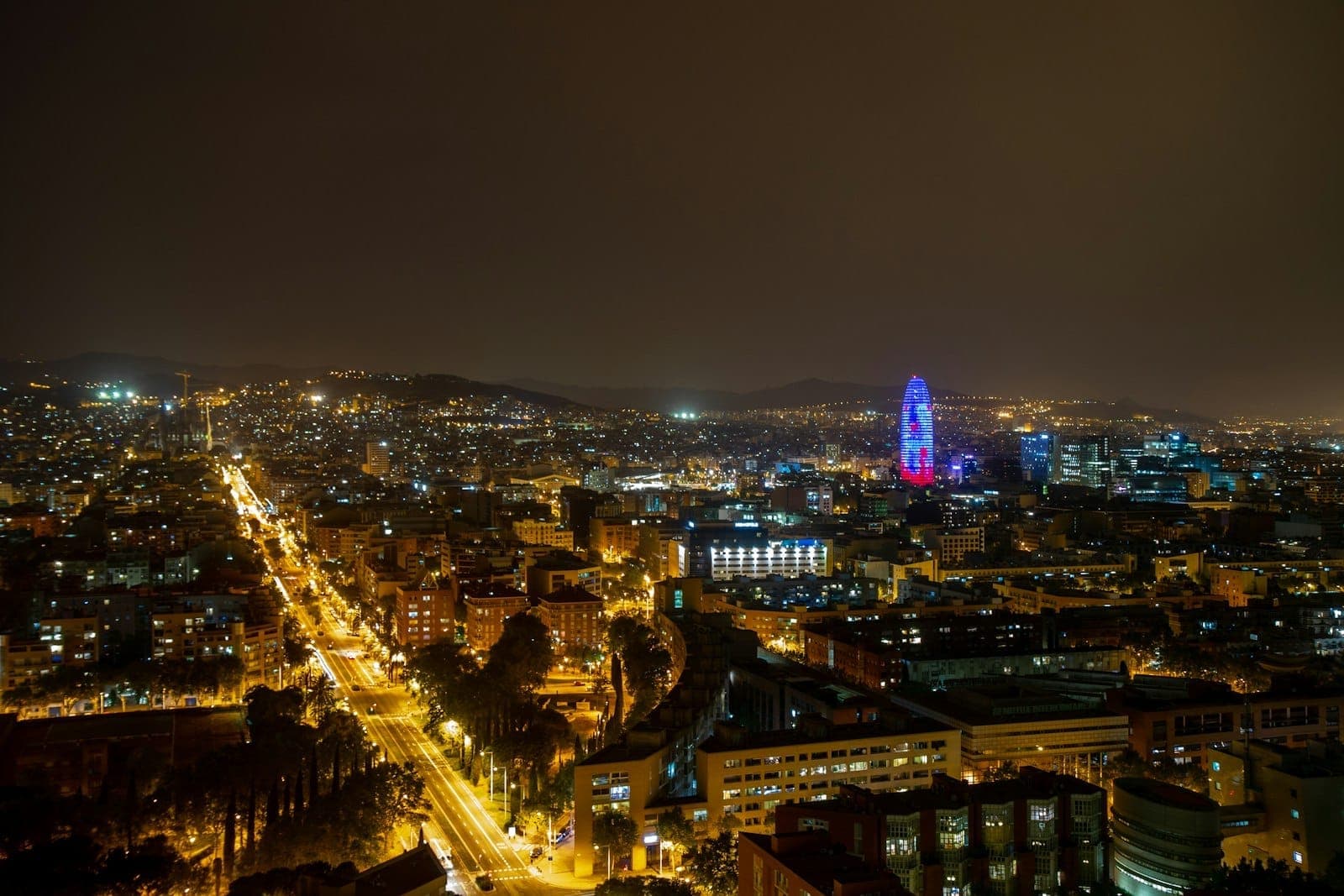Panoramablick bei Nacht über Barcelona mit Stadtlichtern, die sich bis zum Horizont erstrecken, und einem markanten beleuchteten Turm in der Ferne.
