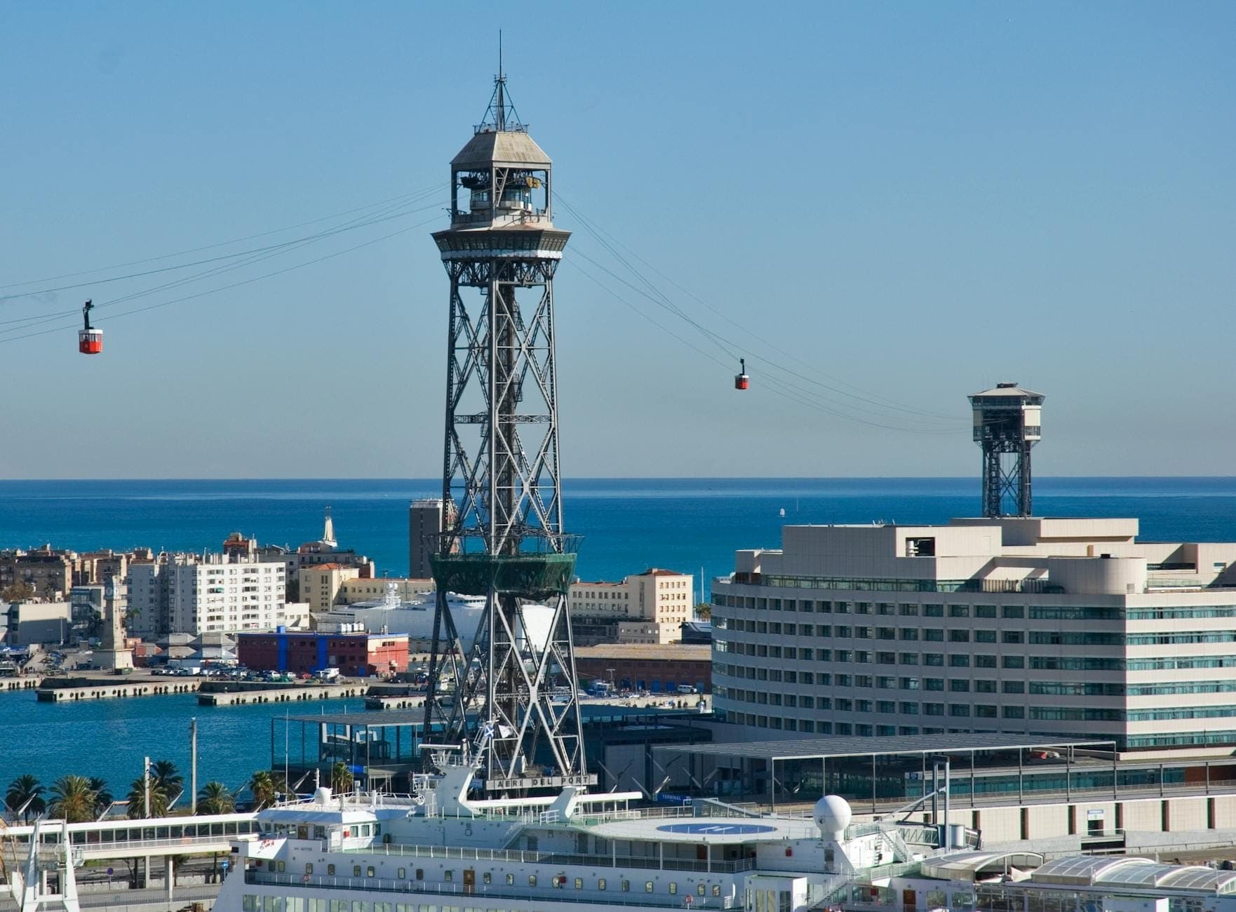Des téléphériques traversent au-dessus du port de Barcelone avec la mer bleue, des bâtiments modernes et la skyline de la ville visibles en contrebas.