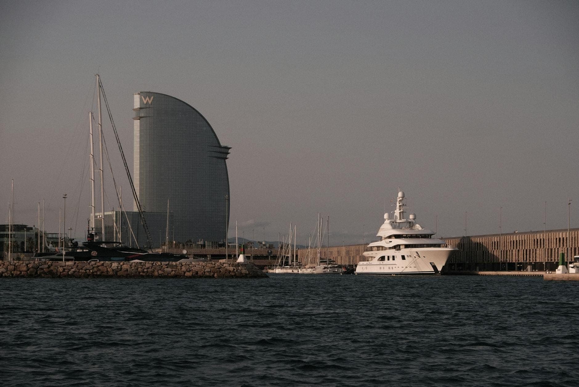Vue en soirée du front de mer de Barcelone avec l'emblématique W Hotel et des yachts amarrés dans la marina, sous un ciel crépusculaire.