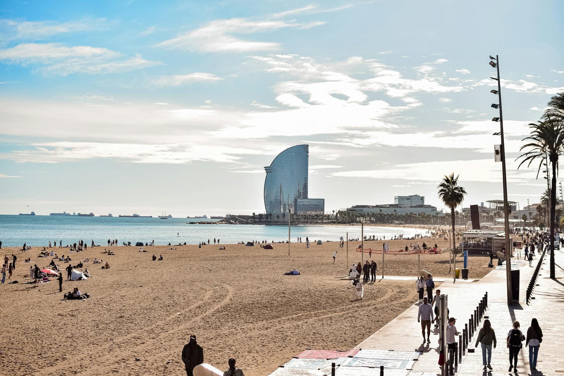 Des personnes qui marchent et font du vélo le long de la promenade de la Barceloneta au bord de la plage, avec le W Hotel et la mer Méditerranée en arrière-plan.