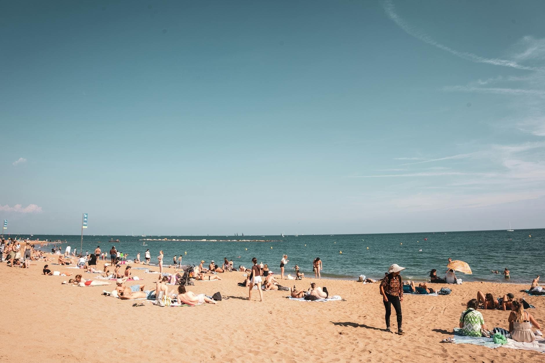 Large plage de sable avec des personnes se relaxant et nageant, peu fréquentée avec une mer calme et un ciel dégagé, ressemblant à la côte plus paisible de Barcelona.
