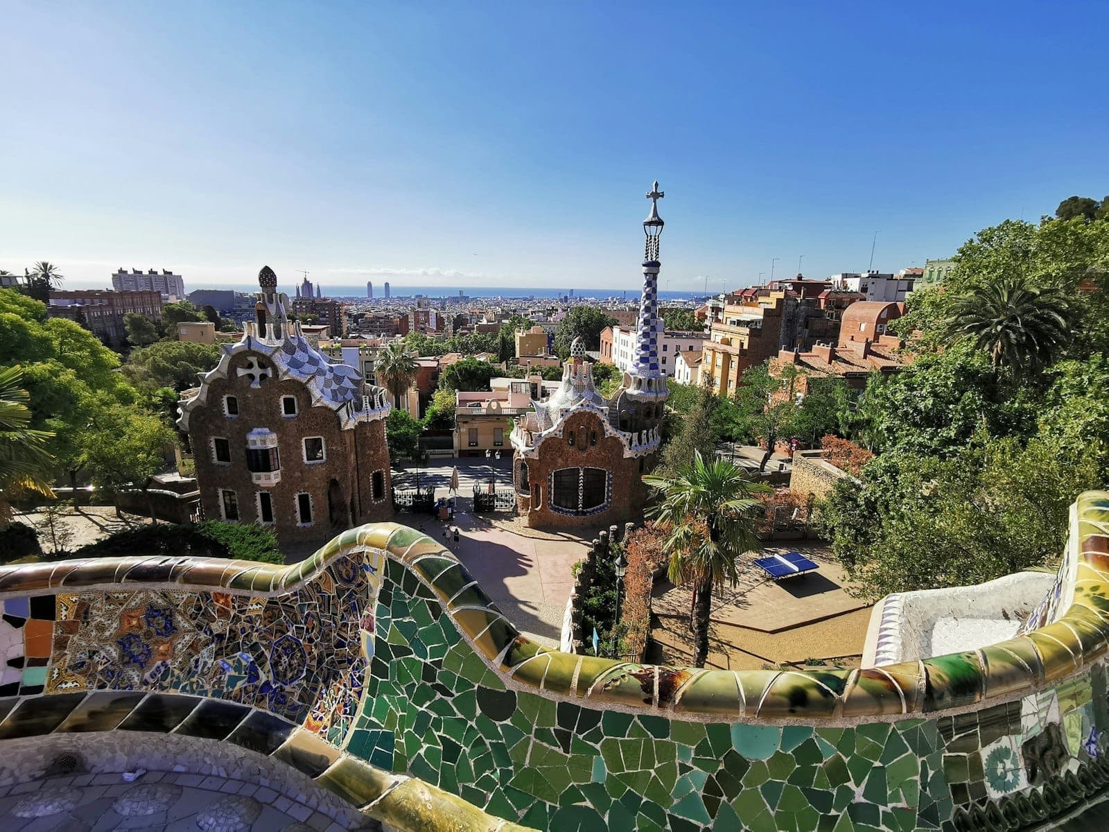 Widok na kolorowy taras z mozaiką Gaudíego w Park Güell i jego fantazyjne domki bramne, z panoramą Barcelony w tle pod bezchmurnym błękitnym niebem.