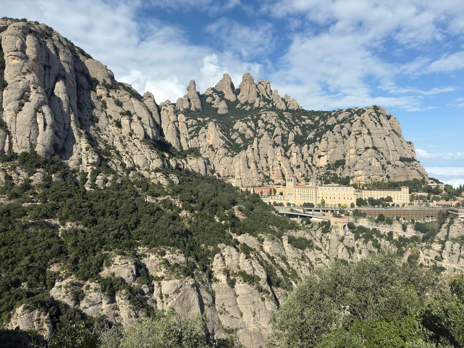 Kısmen bulutlu bir gökyüzünün altında yüksek kayalık dağlara yaslanmış Montserrat manastırının geniş görünümü.
