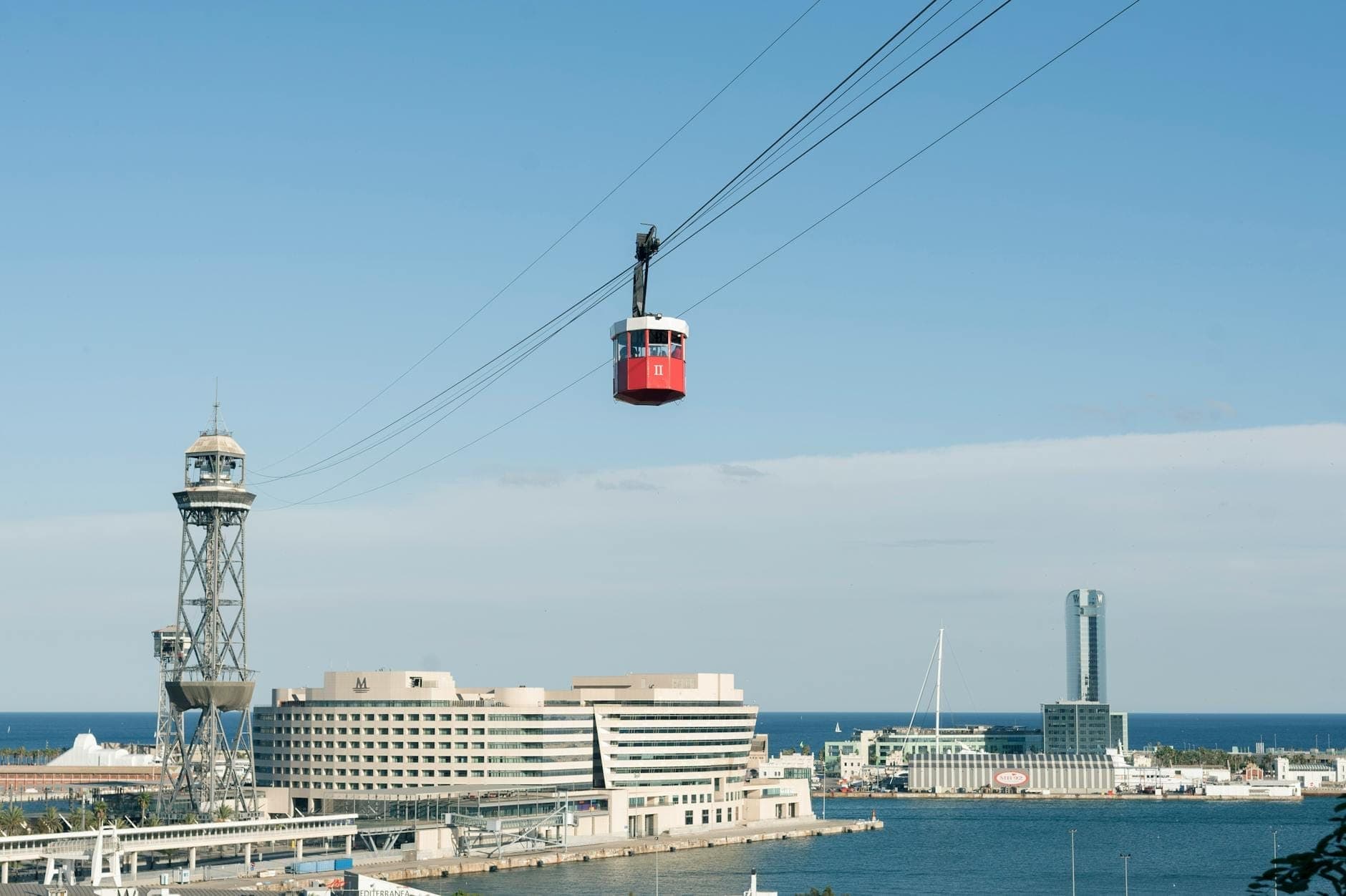 Un teleférico rojo de Montjuïc viaja sobre el paseo marítimo de Barcelona con torres emblemáticas, mar azul y modernos edificios de la ciudad bajo un cielo despejado.