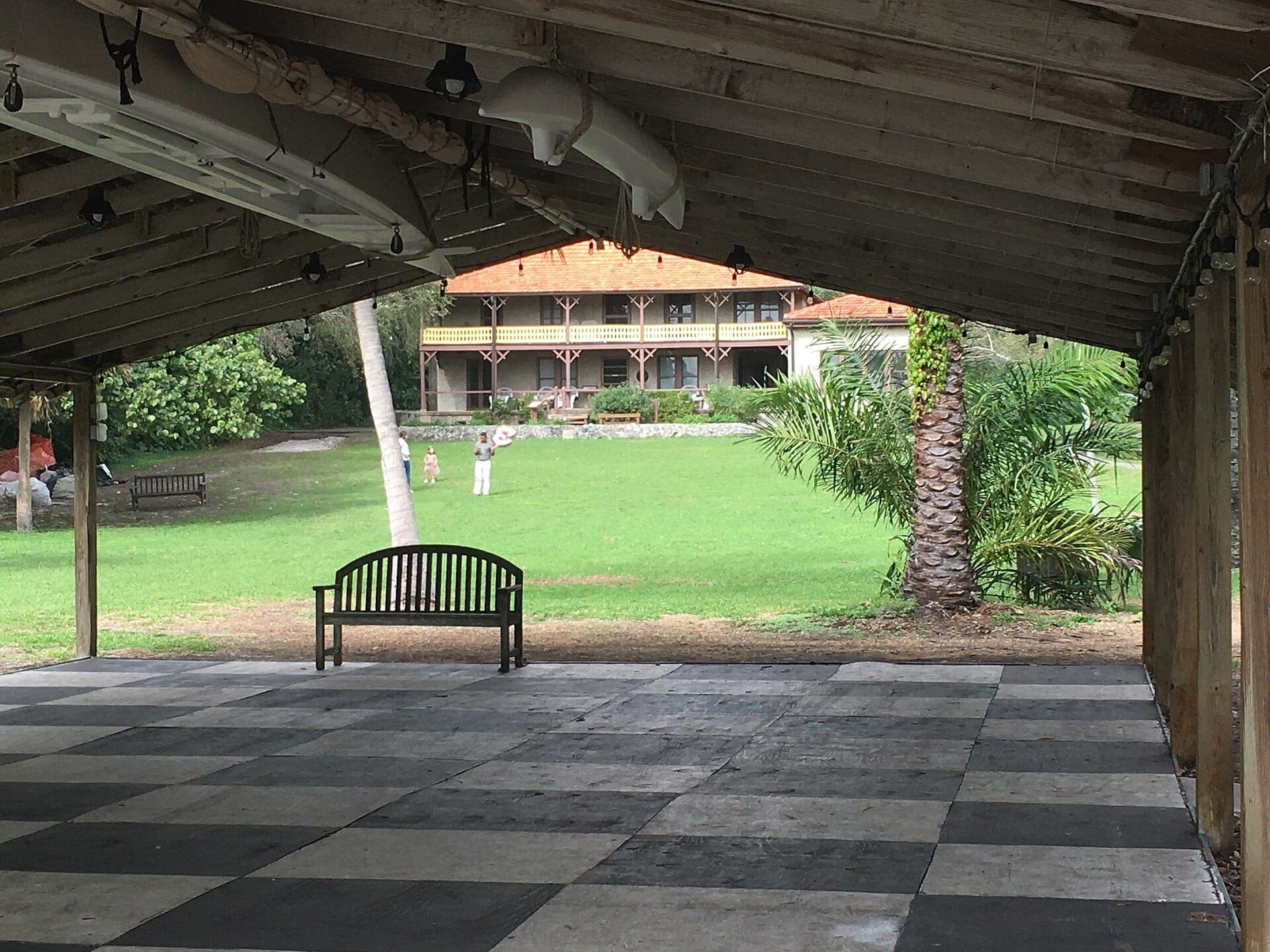 View from a covered pavilion toward Barnacle Historic State Park’s historic house, with open lawn, palm trees, and distant visitors enjoying the sunny grounds.