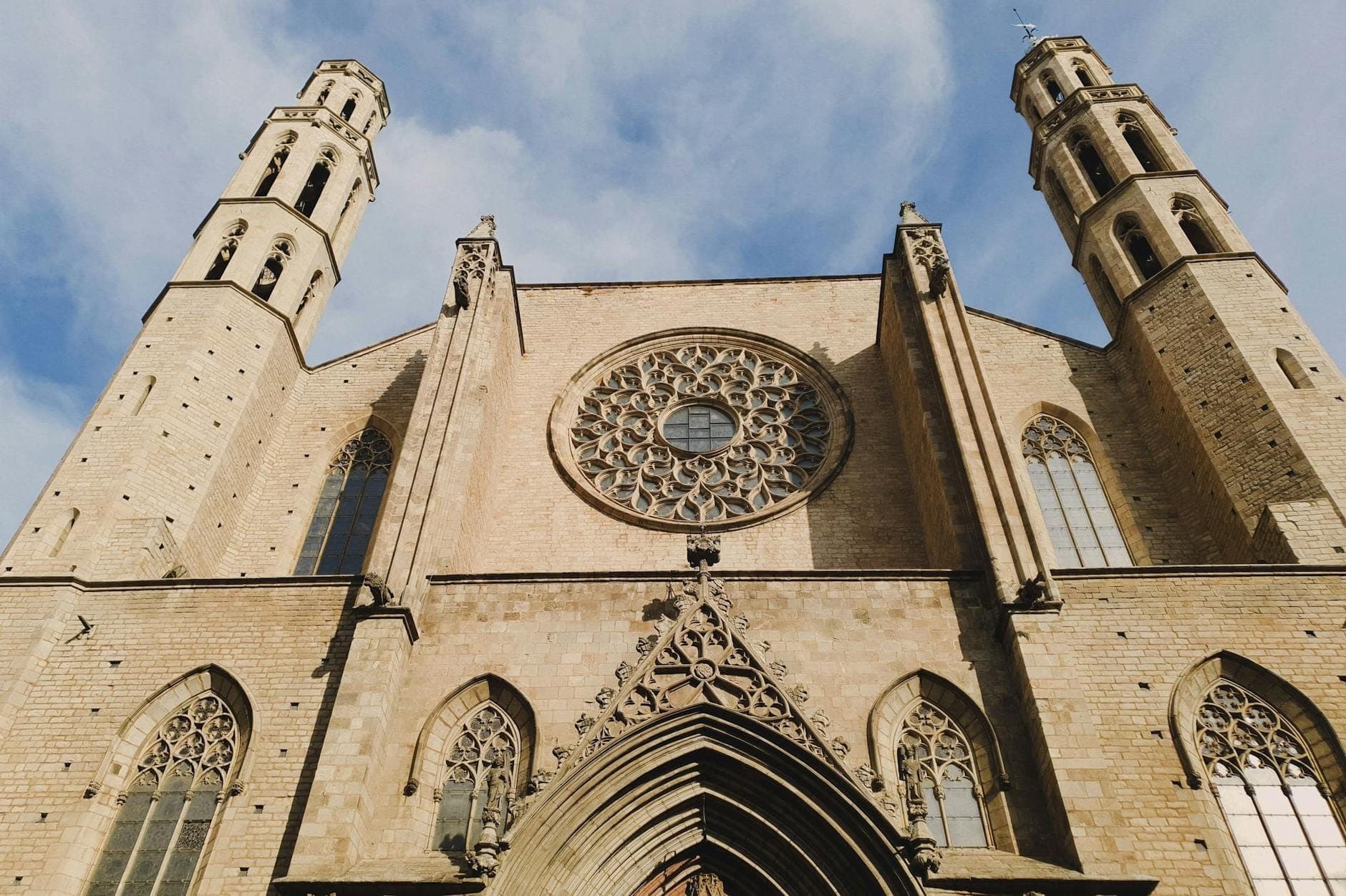 มุมต่ำของด้านหน้ามหาวิหาร Basílica de Santa Maria del Mar ที่เห็นหน้าต่างกุหลาบขนาดใหญ่และหอคอยแปดเหลี่ยมคู่ตัดกับท้องฟ้าสีฟ้า